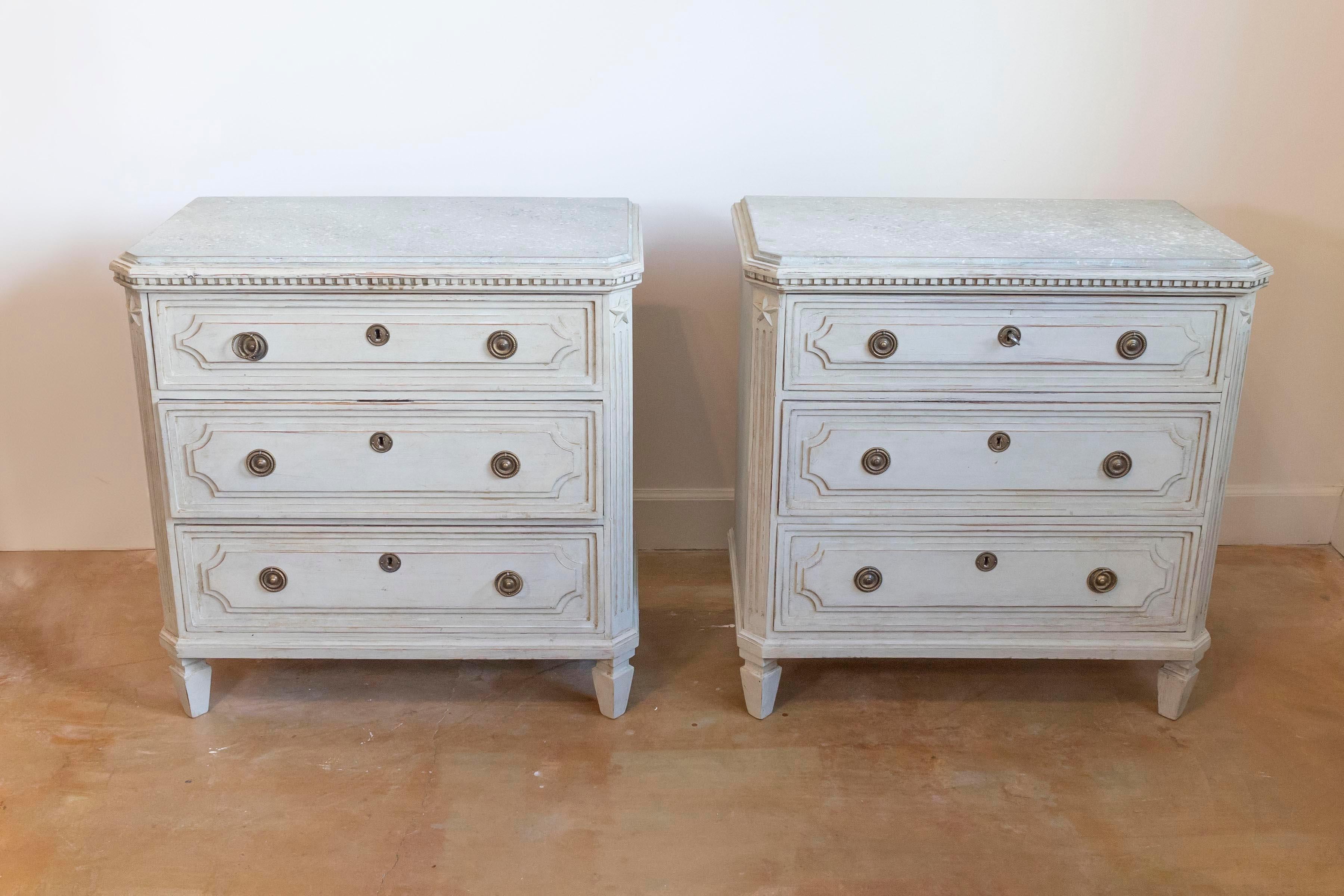 A pair of Swedish Gustavian style painted wood chests from the late 19th century, with marbleized tops, three graduated drawers, dentil molding, carved stars and fluted side posts. Created in Sweden during the last quarter of the 19th century, each