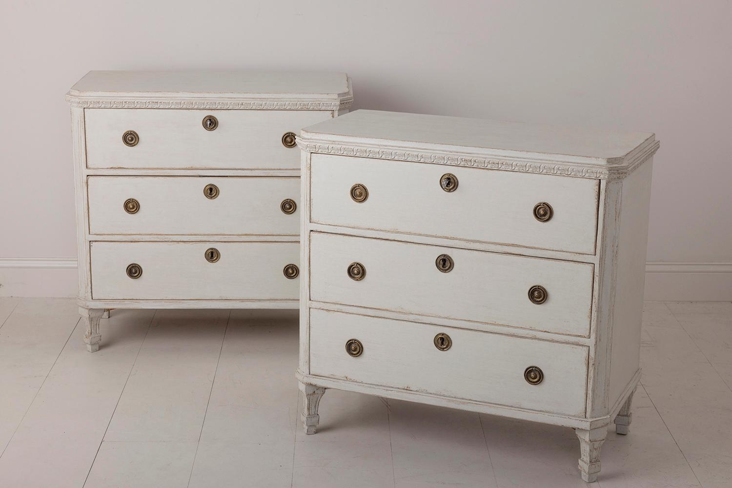 A pair of Swedish commodes in the Gustavian style with carved foliate detail around the tops. Chalky ivory paint. Three large drawers, brass hardware, canted and fluted corner posts, and tapered and fluted legs. Original locks with keys.

 