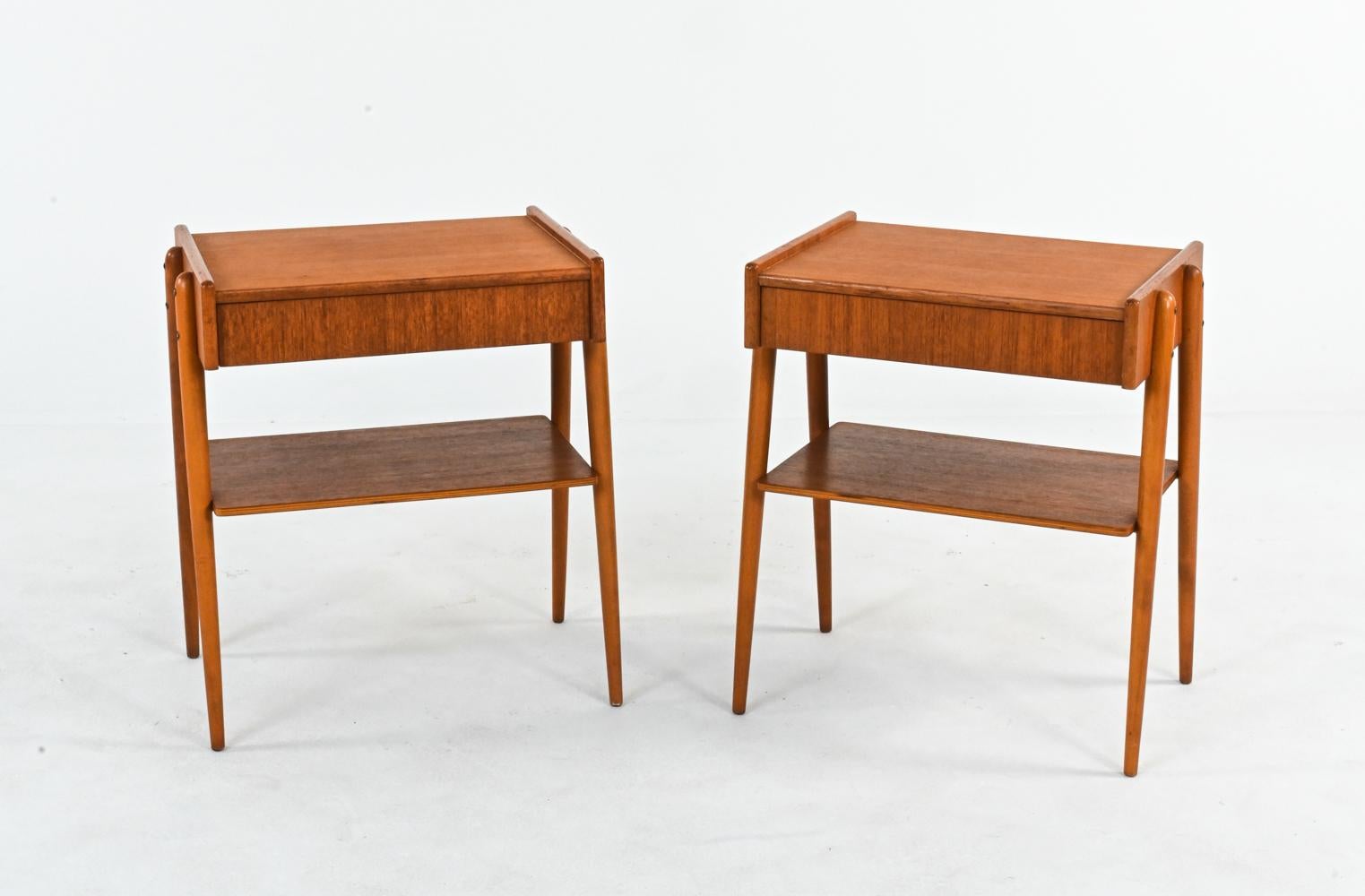 Perfectly proportioned for a small space, this pair of bedside tables by Carlstrom & Co. features warm teak veneers and a classic Mid-Century Modern look. The design consists of a single drawer and shelf suspended between splayed legs, evoking a