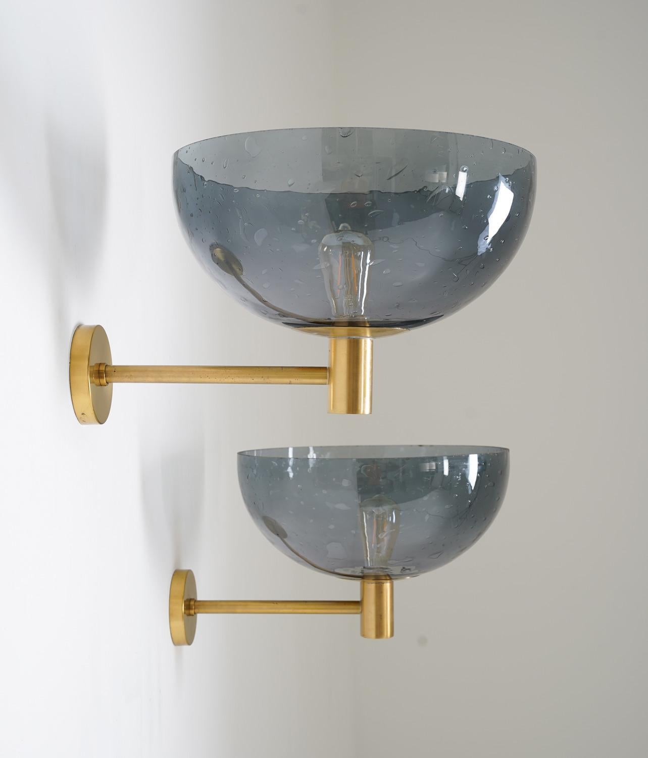 These wall lights are a beautiful example of mid-century Scandinavian design. Manufactured in Sweden in the 1960s, each lamp features a single light source surrounded by a large blue glass shade. The shade is attached to a brass holder, adding to