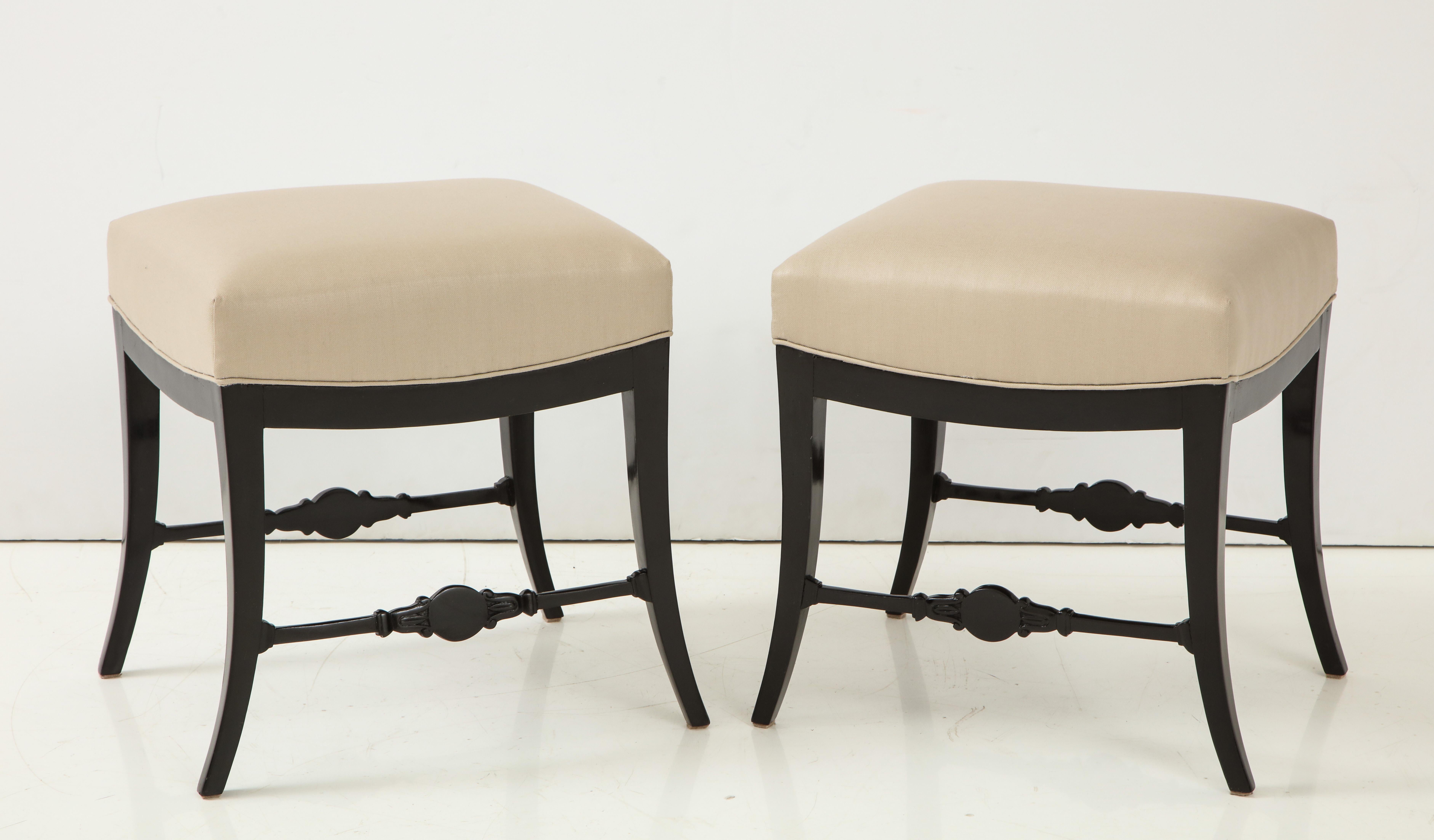 A pair of Swedish neoclassical ebonized and upholstered stools, circa 1830, each with an upholstered seat raised on sabre legs joined by a cross-stretcher with a carved central medallion.