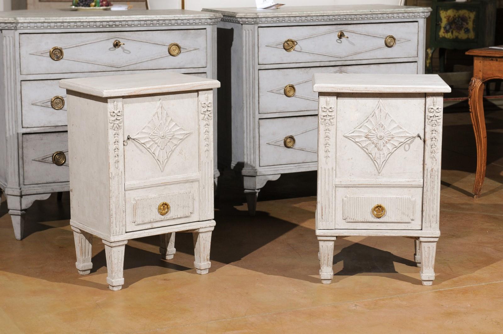 A pair of Swedish Neoclassical style painted nightstand tables from the 20th century, with single doors and carved décor. Born in Sweden, each of this pair of painted nightstand cabinets features a stylish façade presenting a single door adorned