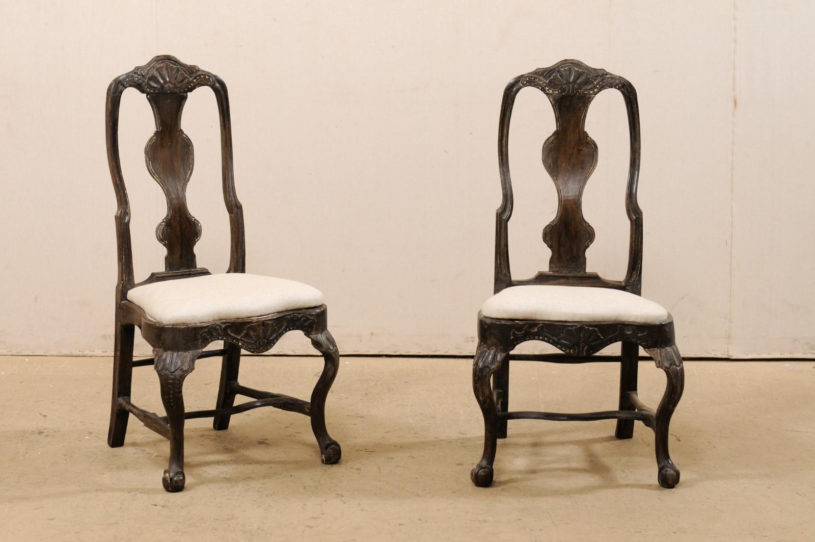 A Swedish pair of period Rococo carved-wood side chairs from the 18th century. This pair of antique chairs from Sweden feature shell motif carvings about the top rail crest and center front seat rail, curvy back splats, stylized knees, and are