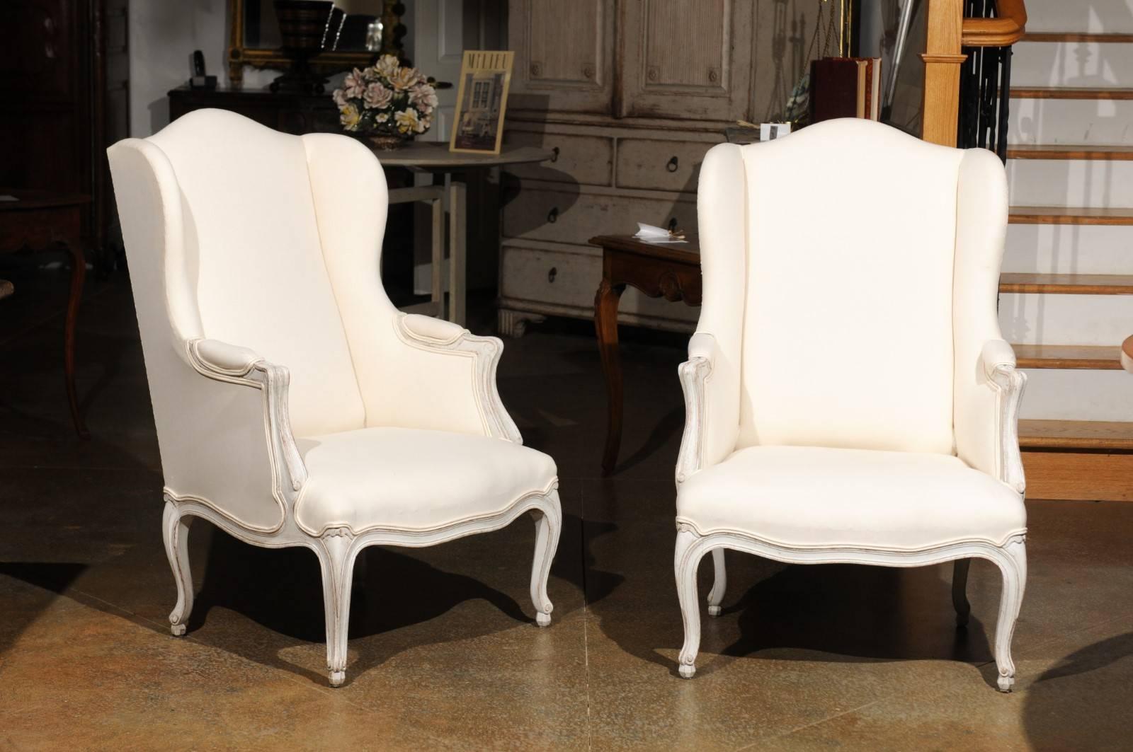 A pair of Swedish Rococo style painted wood wingback bergère armchairs from the early 20th century with new upholstery. Each of this pair of Swedish bergères features a winged camelback, providing both intimacy and protection from drafts. The back