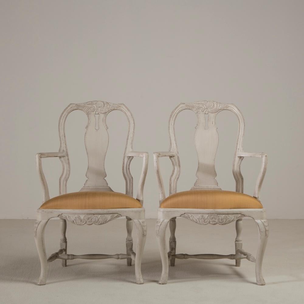 A pair of Swedish Rococo style armchairs in a grey distressed paint. The seat back and base are adorned with foliage, whilst the sabre and cabriole legs have some light carving, circa 1920s.