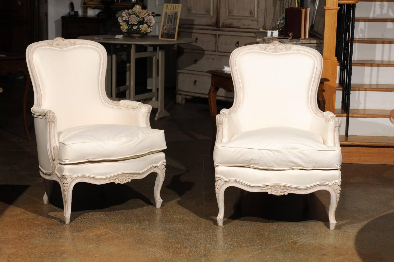 A pair of Swedish painted Rococo style bergères chairs from the late 19th century, with carved crests, scrolled arms, cabriole legs and upholstery. Each of this pair of Swedish bergères features a curved back, topped with a carved floral crest. This