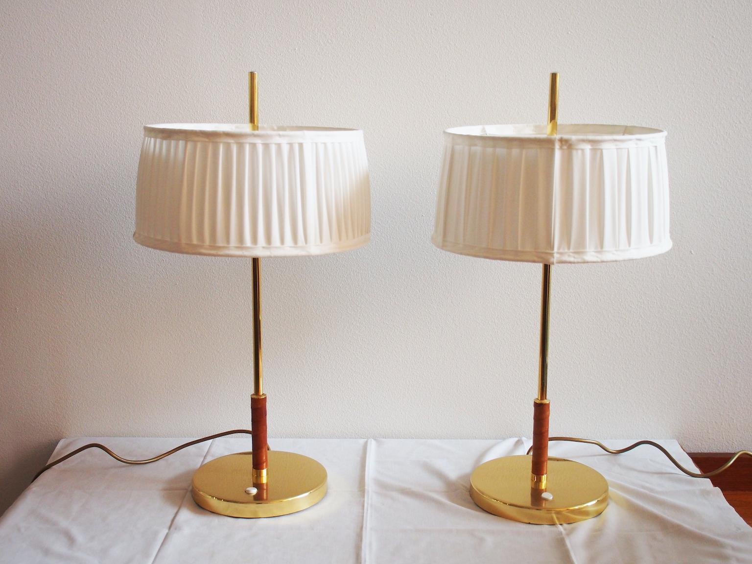 Paire de lampes de table en laiton avec détails en cuir brun. Fabriqué en Suède par Örsjö Industri. Abats-jour en tissu blanc légèrement différents.