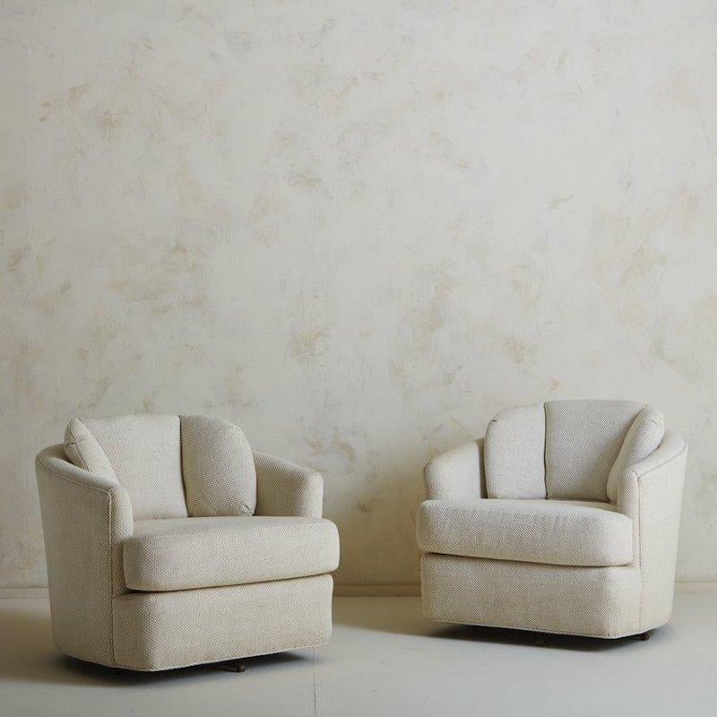 A pair of midcentury swivel lounge chairs by Thayer Coggin featuring rounded barrel frames and removable tri-fold back cushions. These chairs were reupholstered by previous owners in a textural taupe and cream fabric. They have metal star swivel