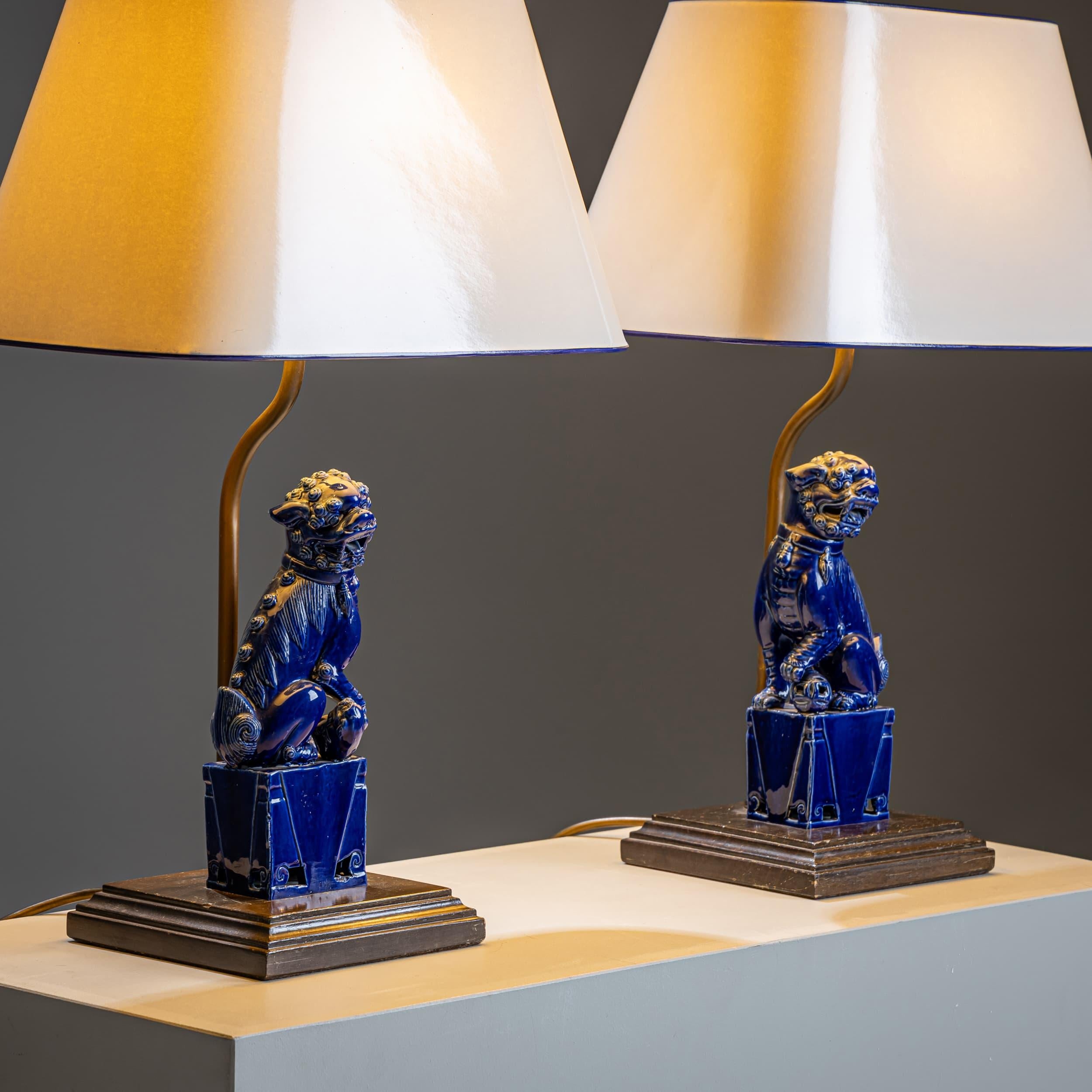 Pareja decorativa de lámparas de mesa con las tradicionales figuras chinas del perro Fu como base, fabricadas en cerámica con un llamativo esmalte azul cobalto. Cada perro Fu se alza sobre un pedestal estilizado y está coronado por una pantalla