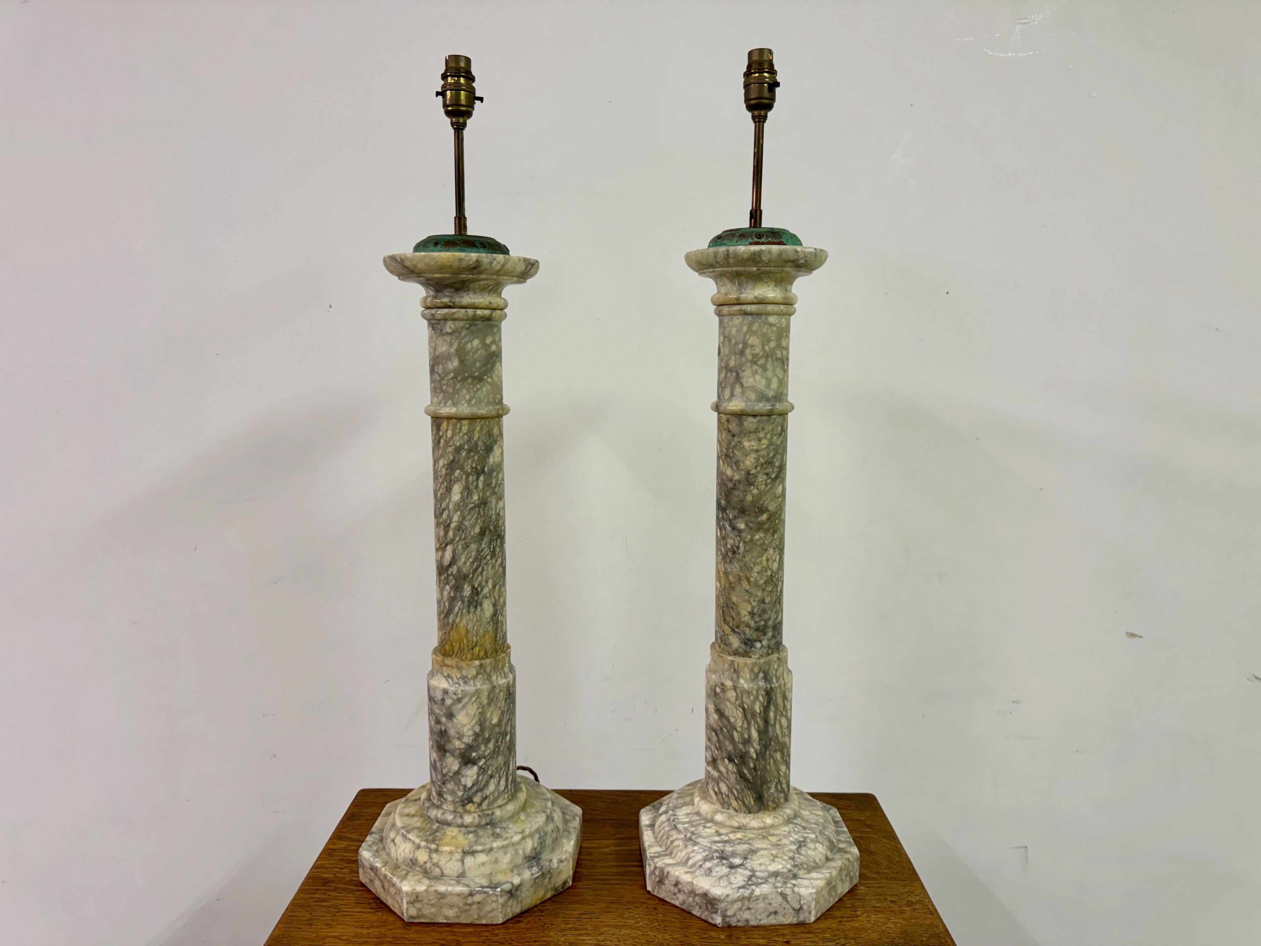 This pair of table lamps are crafted from alabaster, a material known for its fine grain and translucency, often used in decorative objects and lighting. The lamps exhibit a classical columnar design, featuring tall, slender shafts with subtle