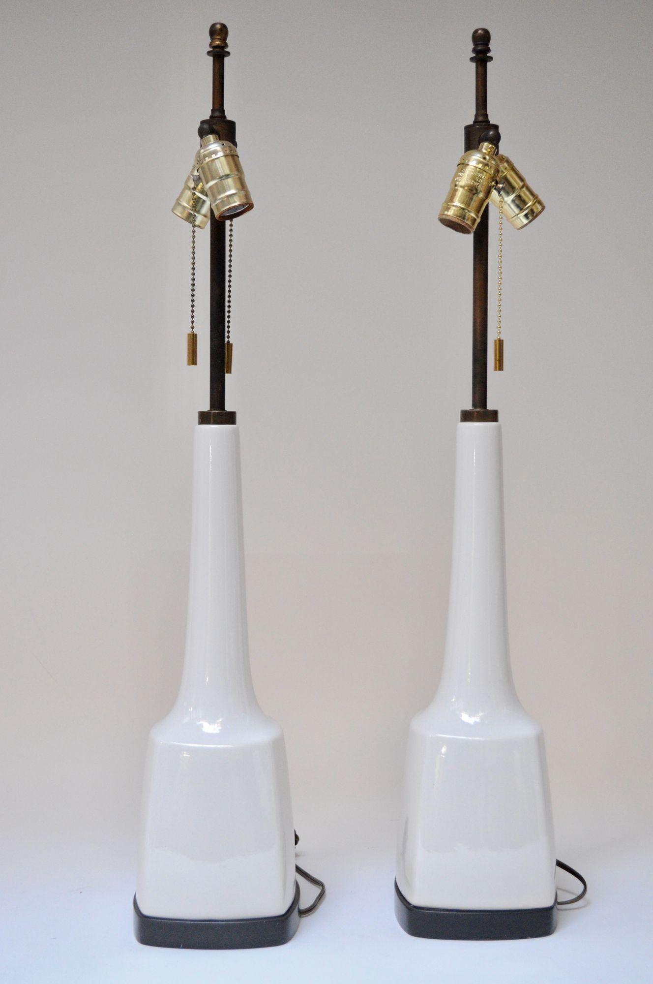 Pair of Mid-Century American Modern tables lamps (ca. 1950s, USA). Composed of patinated brass stems and white porcelain forms, all supported by square ebonized wooden platform bases. Along with the wiring, the dual brass sockets and on/off pull