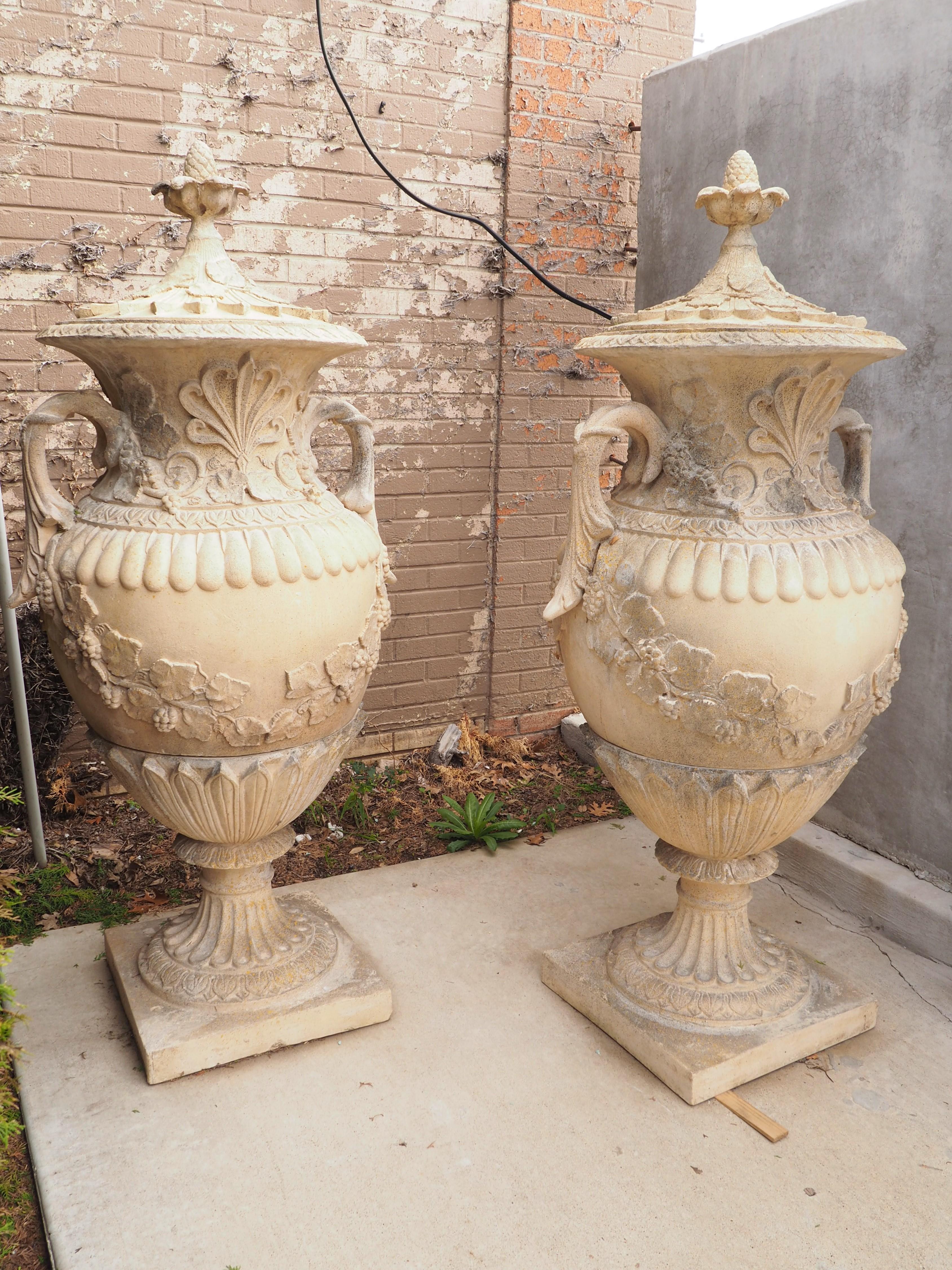 This beautiful pair of French urns, measure just over 5 feet tall. Each urn consists of three pieces of cast stone, all with nature-inspired motifs. Both lids are topped with a pinecone finial emerging from a curled leaf cup above two layers of