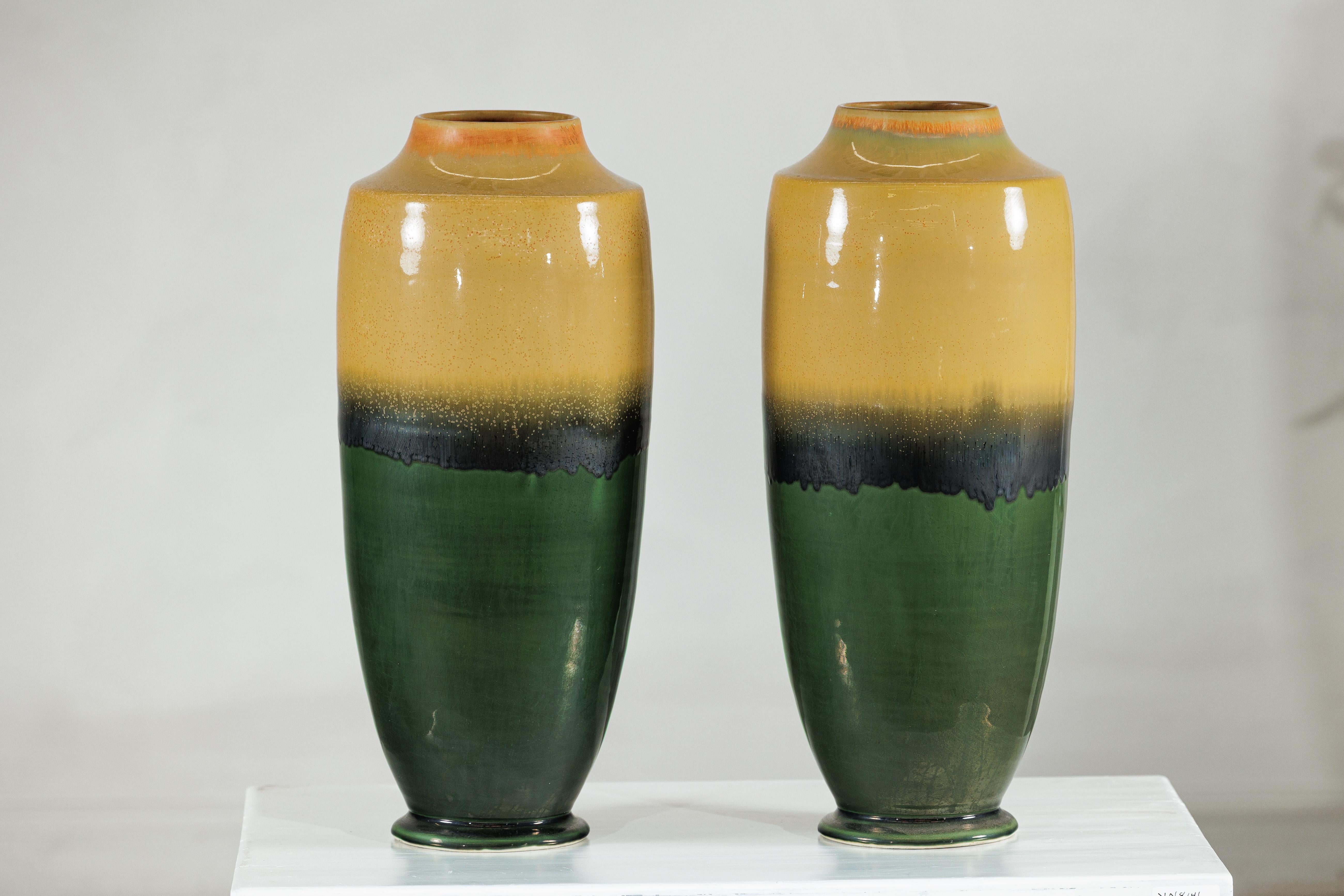 Questa sorprendente coppia di vasi alti in ceramica mette in mostra l'abilità della smaltatura a strati, con una tavolozza che si muove senza soluzione di continuità dal senape terroso attraverso una fascia fumosa di nero fino a un verde intenso e