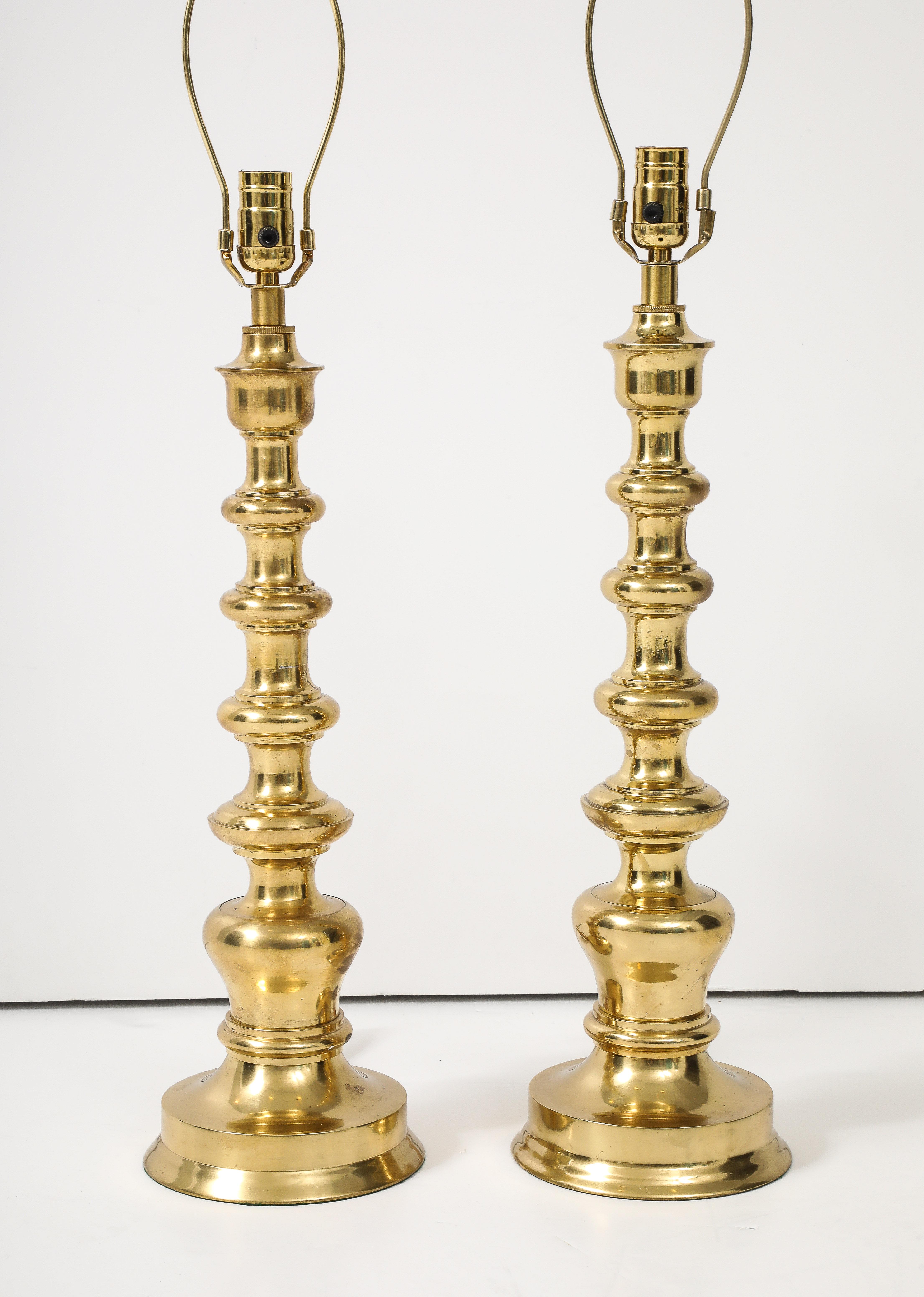 Ein auffälliges Paar hoher Messinglampen in zylindrischer Form mit leicht abgestuften Ringen, die in einem runden Messingsockel enden.  Die elegante Silhouette der Lampen passt sowohl in ein traditionelles als auch in ein modernes Ambiente.