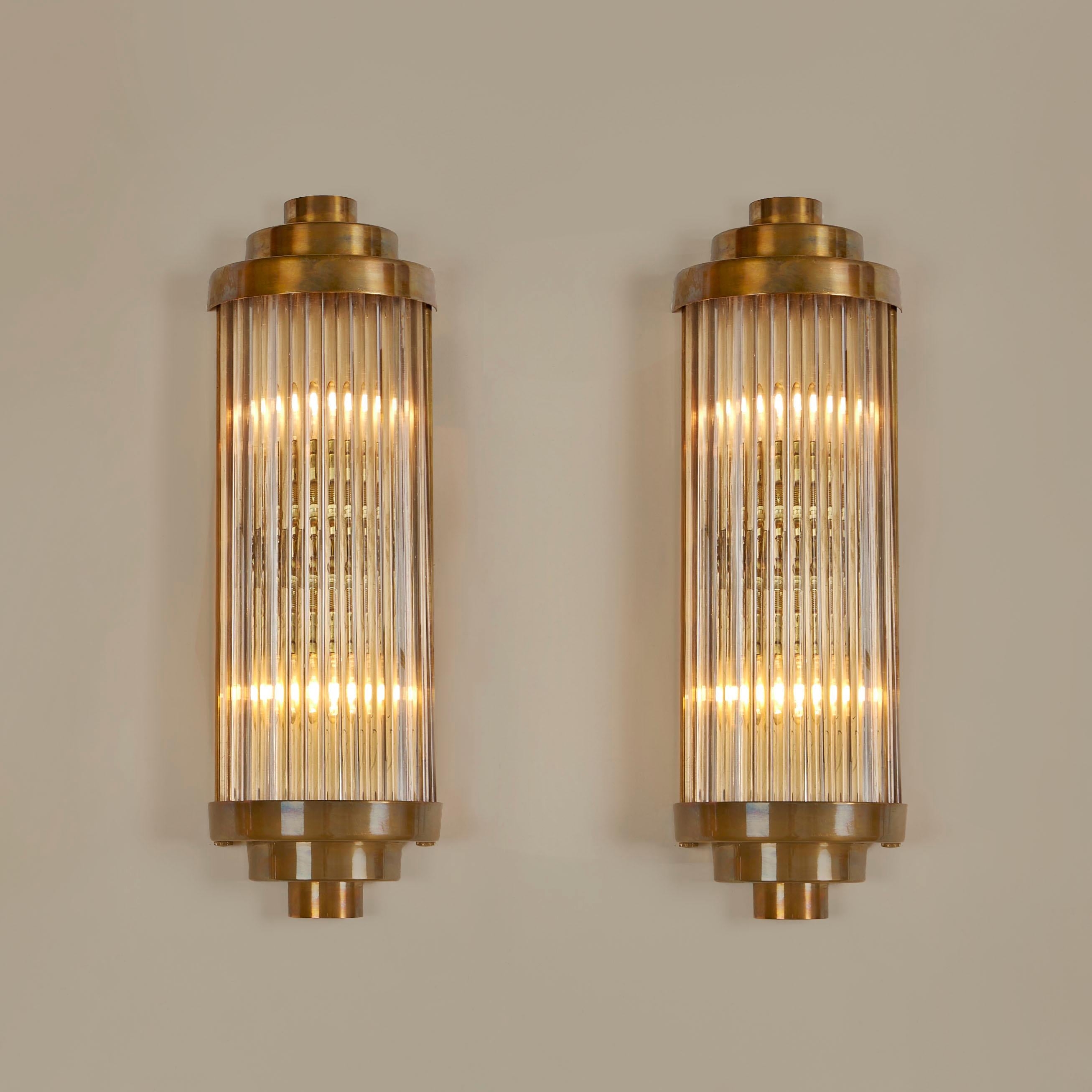These ‘Ravello’ wall lights takes its inspiration from a timelessly classic Art Deco design.
Multiple Italian glass rods form a semi-circle which is capped, top and bottom by 3 tiers of curved brass. The brass back plate holds two light bulbs and