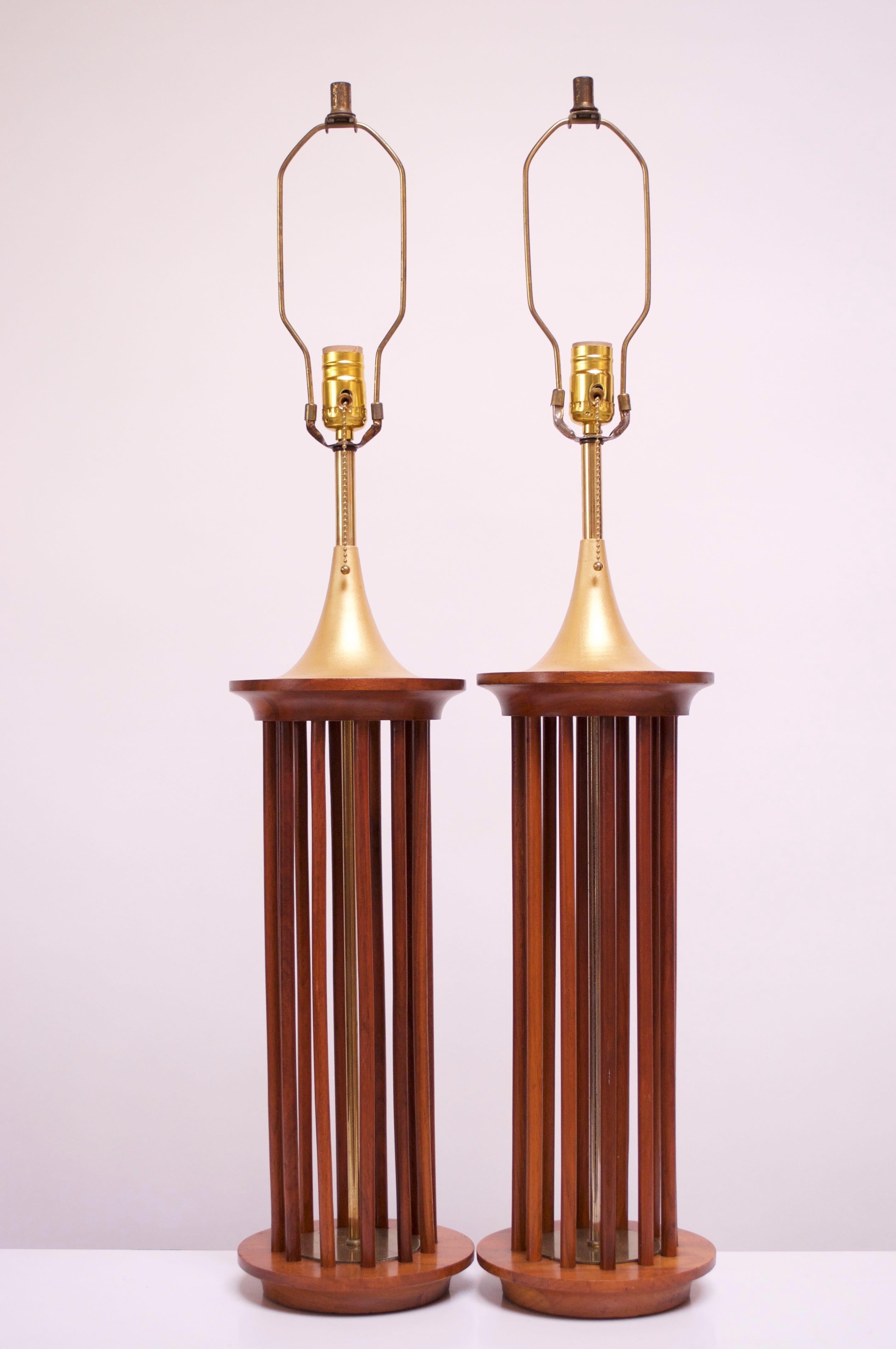 Elegant pair of 1950s American Modern table lamps composed of a solid walnut platform base and spindle-constructed frame that houses an inset brass circular base and brass-plated rod at the center. Lamps are finished on the top by gilt aluminum