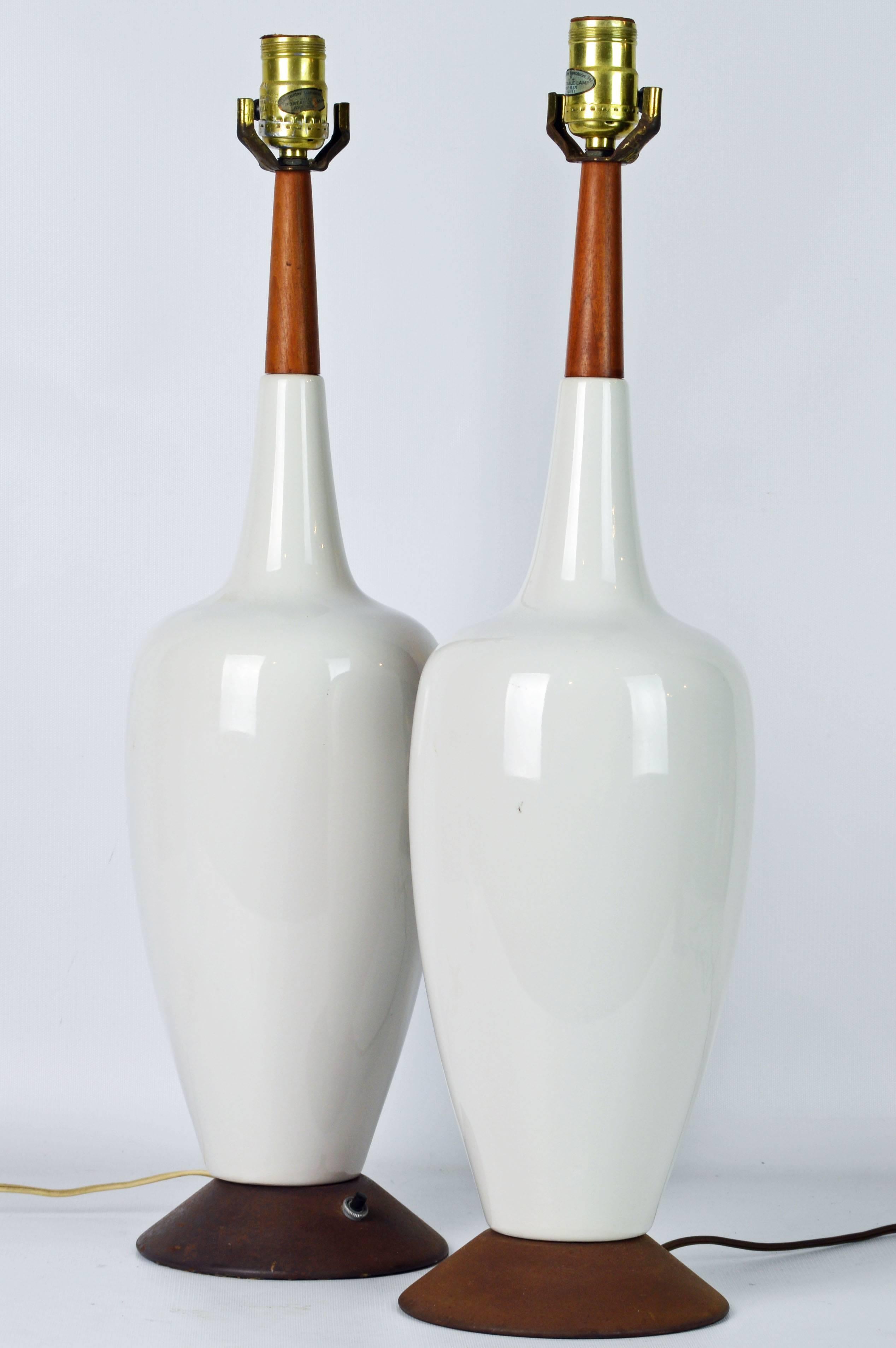 Standing 24.5 inches tall without harps and shades Danish midcentury style table lamps make a fine classic modern statement. The main ceramic white glazed amphora shaped bodies are topped by necks of teak and resting on oxidized circular bases.