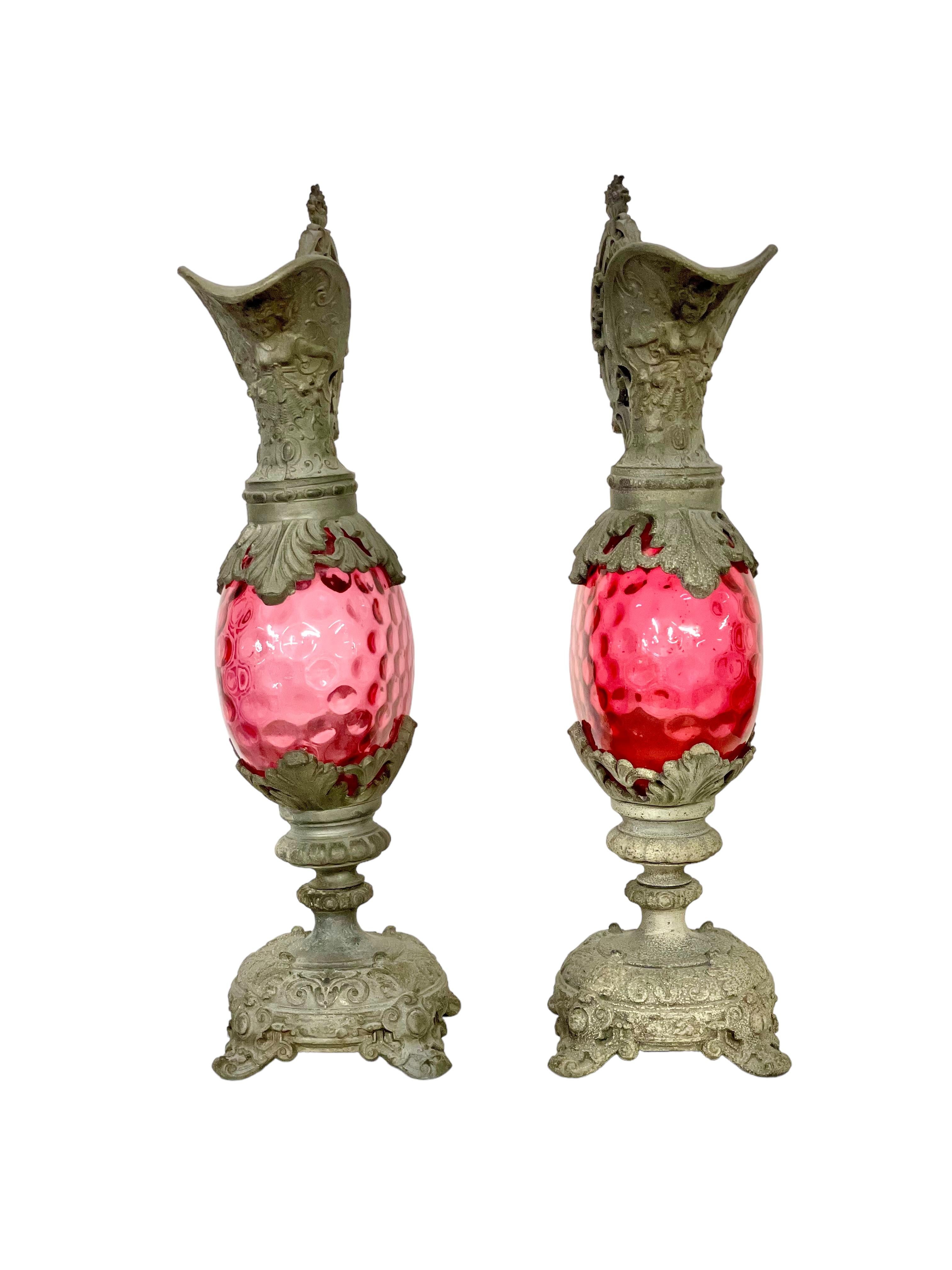 A most elegant pair of Napoleon III period decorative mantle ewers, exquisitely crafted from silver-plated metal with globe-shaped reservoirs of cranberry-coloured glass. These delightful mid-19th century jugs feature extremely ornate high handles,
