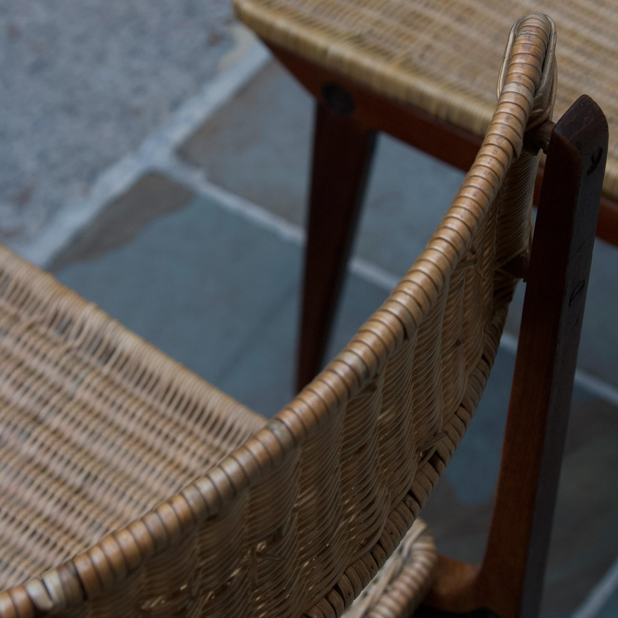 A pair of teak dining chairs, likely to have been made in France during the early 1950s. 

The seat and back of this pair are wicker, woven around a steel frame which is fixed to the teak body of the chair. The curved wooden body is formed by
