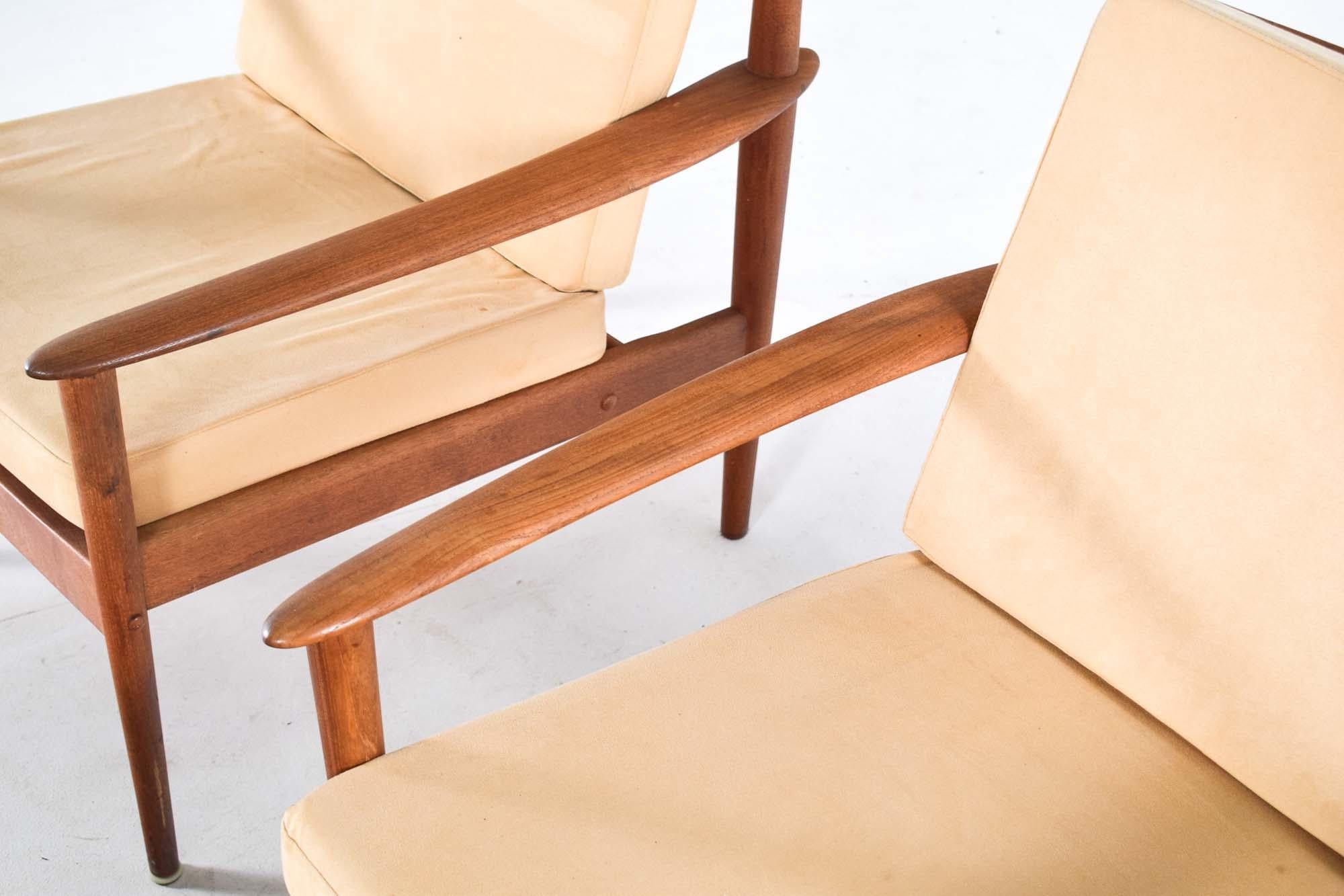 Cette image présente une paire de fauteuils modernes du milieu du siècle accompagnés d'un tabouret assorti, conçus par Grete Jalk. Les chaises et le tabouret sont fabriqués en bois de teck, qui a vieilli pour prendre une teinte chaude et mielleuse,