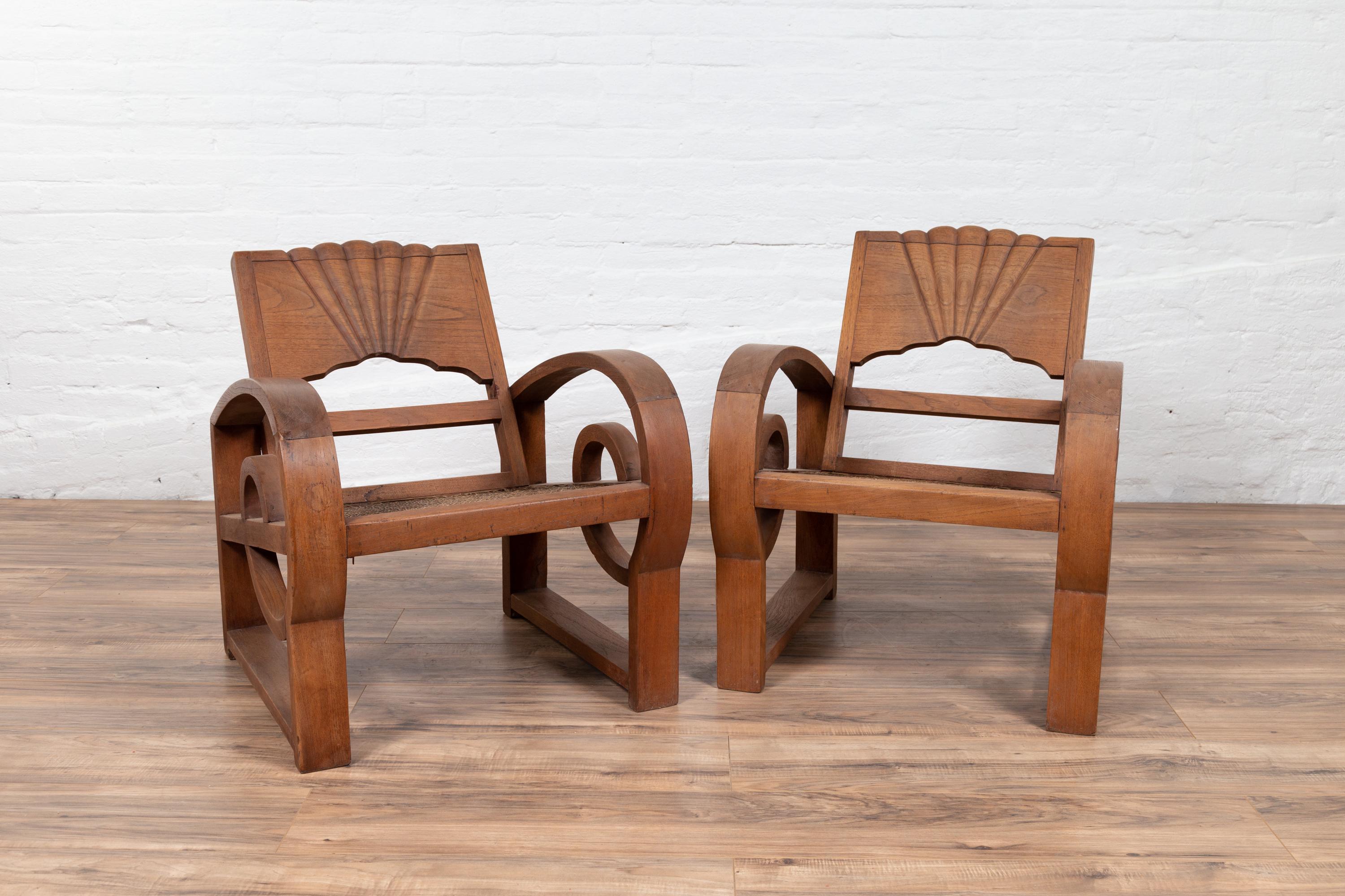 A pair of antique Indonesian teak wood country armchairs from Madura with rattan seats and looping arms. Born on the island of Madura off of the northeastern coast of Java, this charming pair of early 20th century armchairs each features a slightly