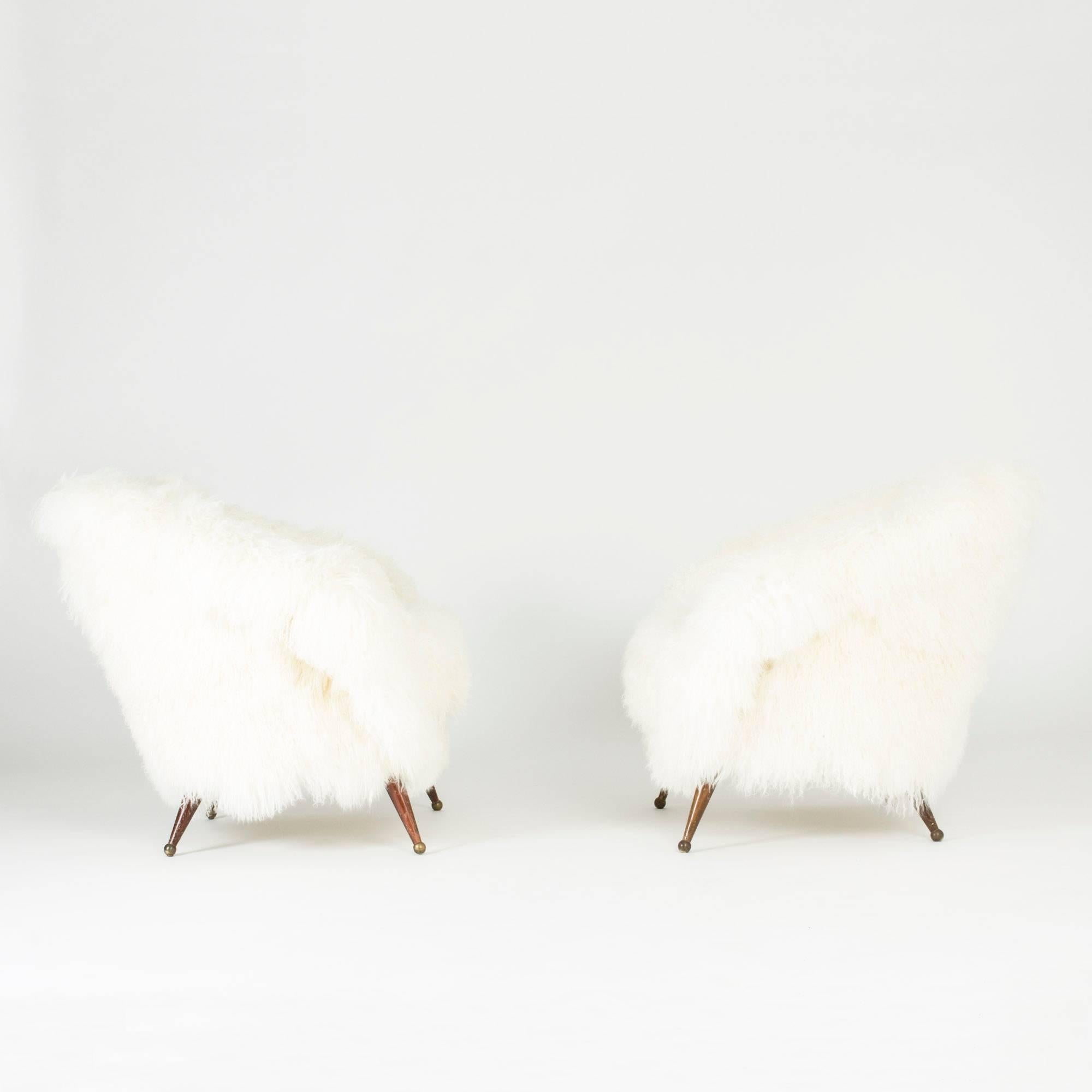 Pair of stunning “Tellus” lounge chairs by Folke Jansson, upholstered with white, long, wavy sheepskin. Rounded, wide and low design with slender legs with brass balls as feet.