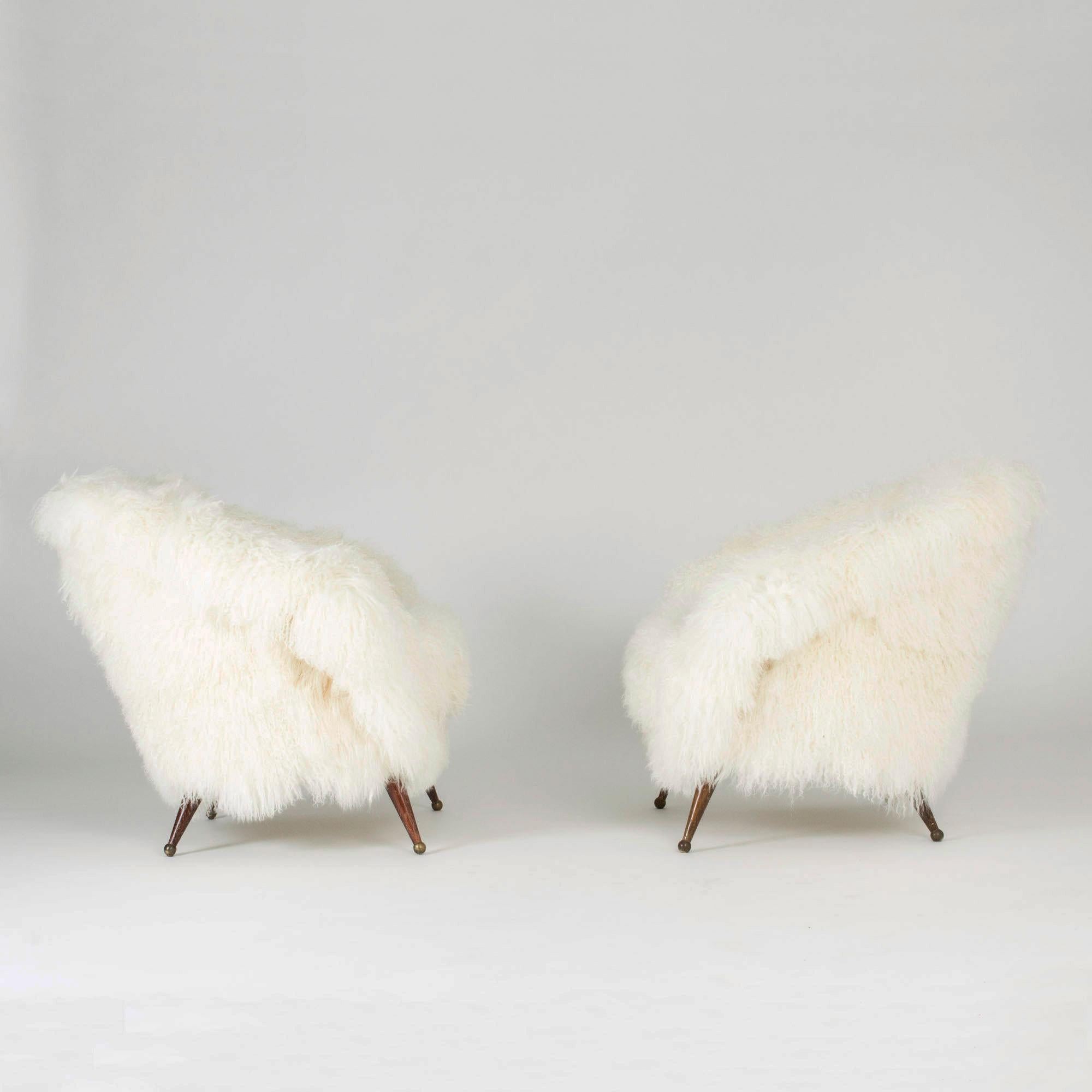 Pair of stunning “Tellus” lounge chairs by Folke Jansson, upholstered with white, long, wavy Tibetan sheepskin. Rounded, wide and low design with slender legs with brass balls as feet.