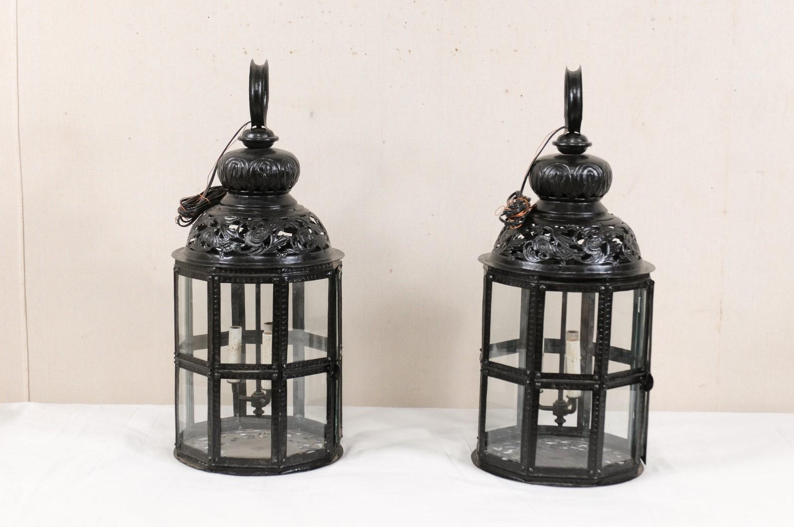 A pair of three-light black metal hanging Moroccan inspired lanterns. This pair of lanterns from Europe have an octagonal-shaped body, within a rounded top and bottom piece. The frame is black metal with glass set within the panels. There is a domed