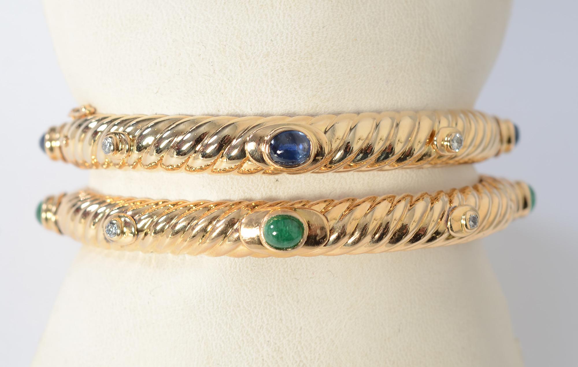 A classic pair of fluted gold bangle bracelets by Tiffany. They are enhanced with diamonds and cabochon emeralds on one; diamonds with sapphires on the other.
The bracelets are hinged with safety clasps. Inside diameters are 2 3/16