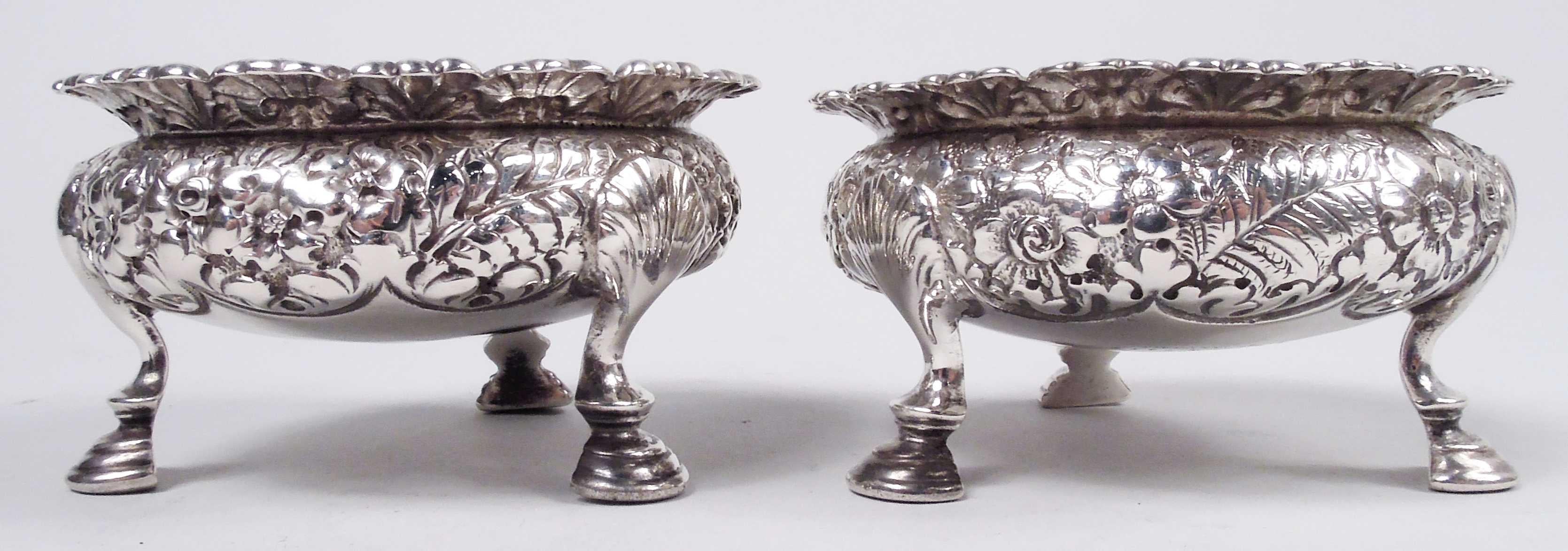Pair of Victorian Classical sterling silver open salts. Made by Tiffany & Co. in New York. Each: Bellied bowl on 3 shell-mounted hoof supports. Repousse flowers and fronds. Everted rim with repousse scallop shells. Interlaced script monogram