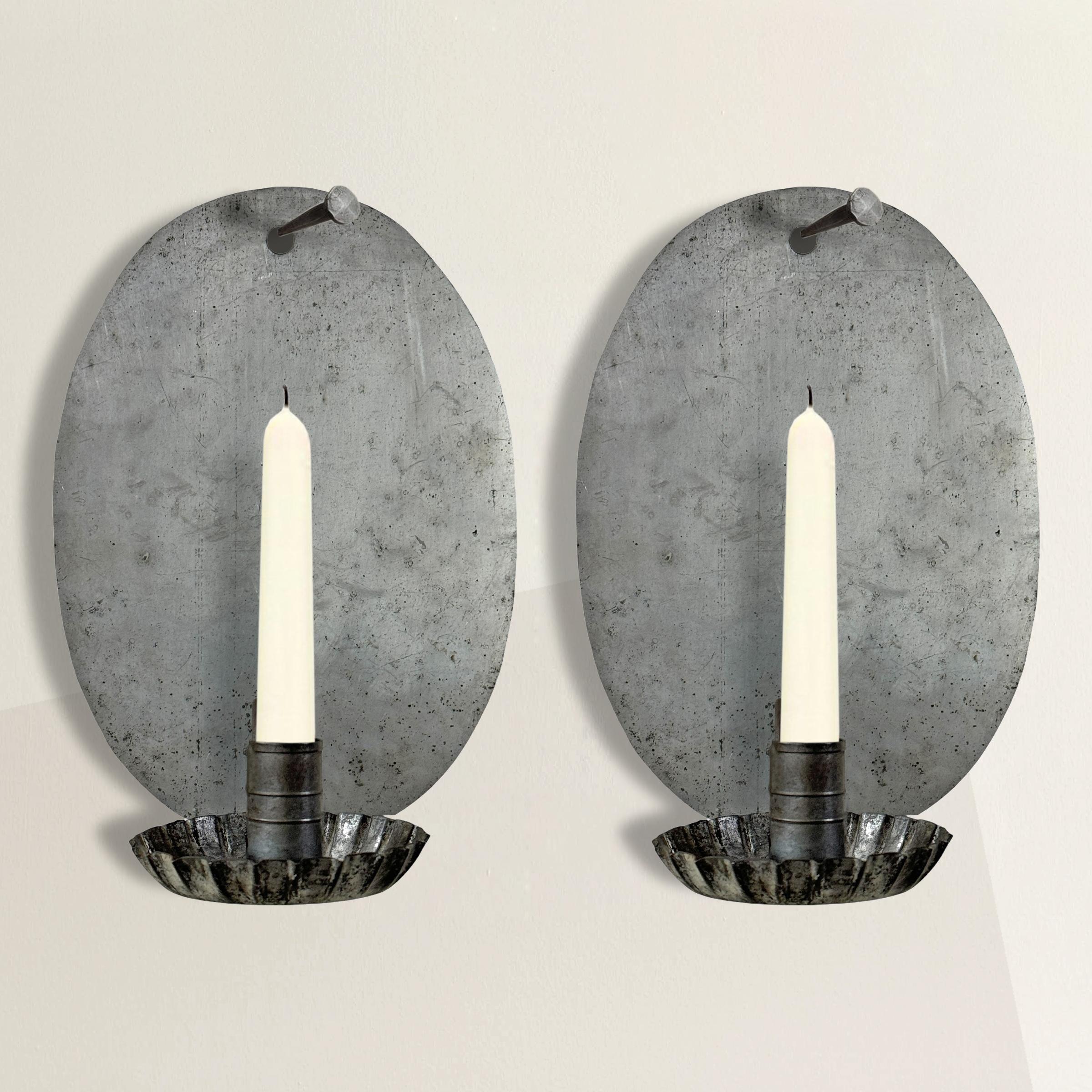 This pair of late 20th-century American tin candle sconces combines modern craftsmanship with a nostalgic nod to early American design. Each sconce features a large concave oval back plate, thoughtfully designed to reflect and enhance the glow of