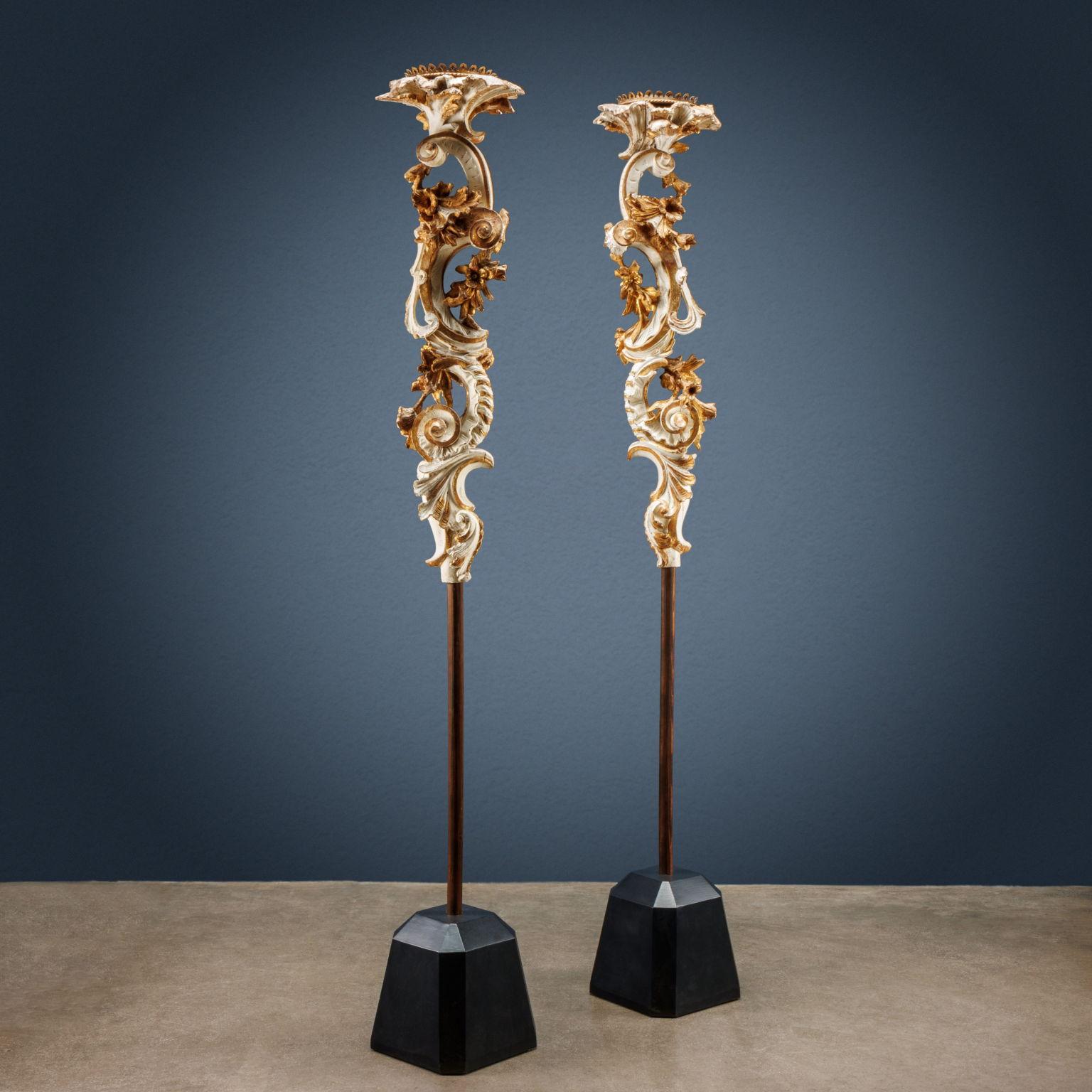 Pair of lamp-holders in carved pine wood, lacquered white and partially gilded, supported by a cylindrical shaft. Composed of large opposing “C” volutes, they are carved with curls and leaf motifs, embellished by a flowering branch that descends