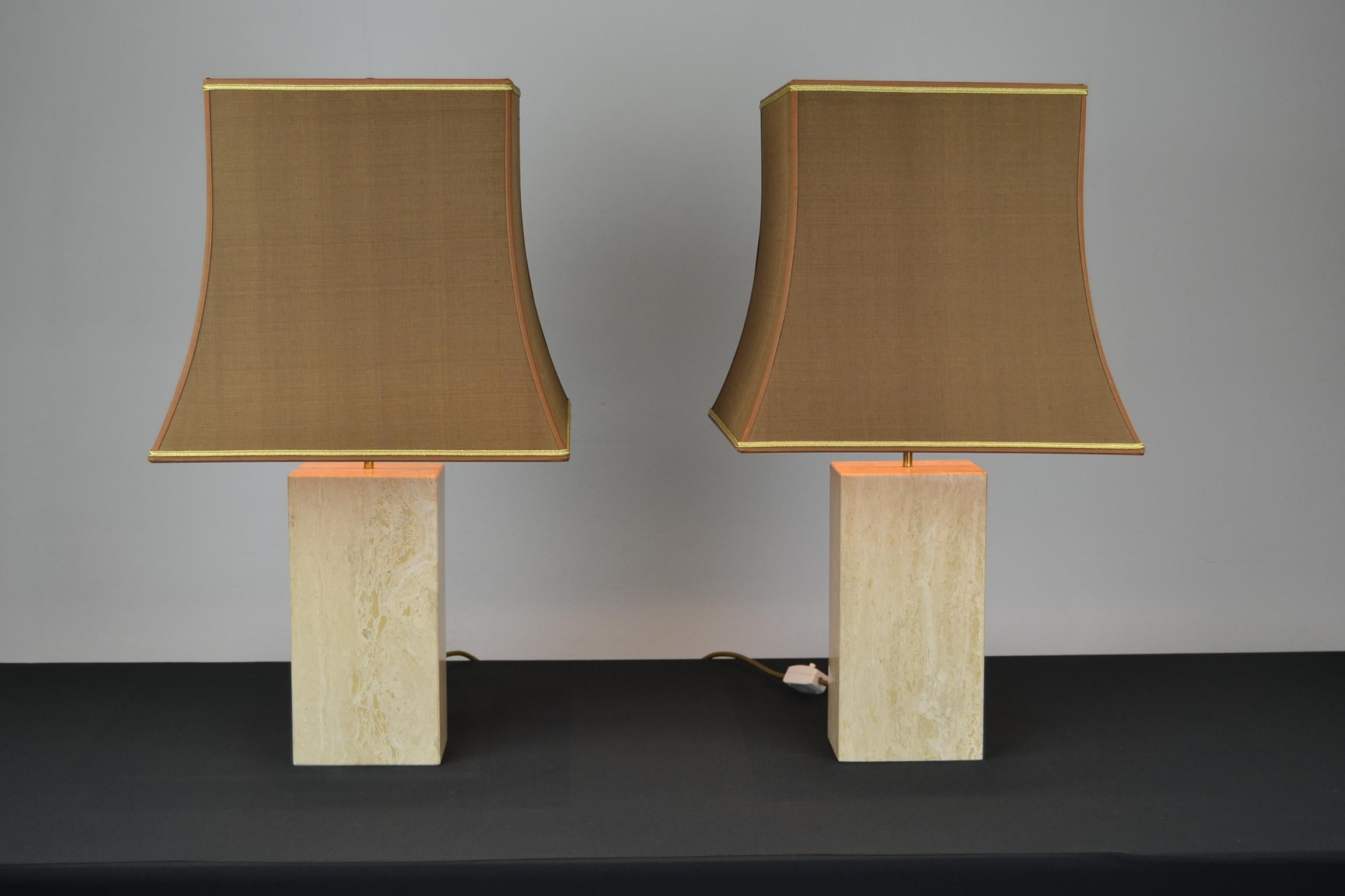 Impressive pair of travertine table lamps from the 1970s.
These square columned table lamps both have their handmade brown shades with gold edging and gold color inside what reflects the light. They have each 2 bulbs which are connected by brass to