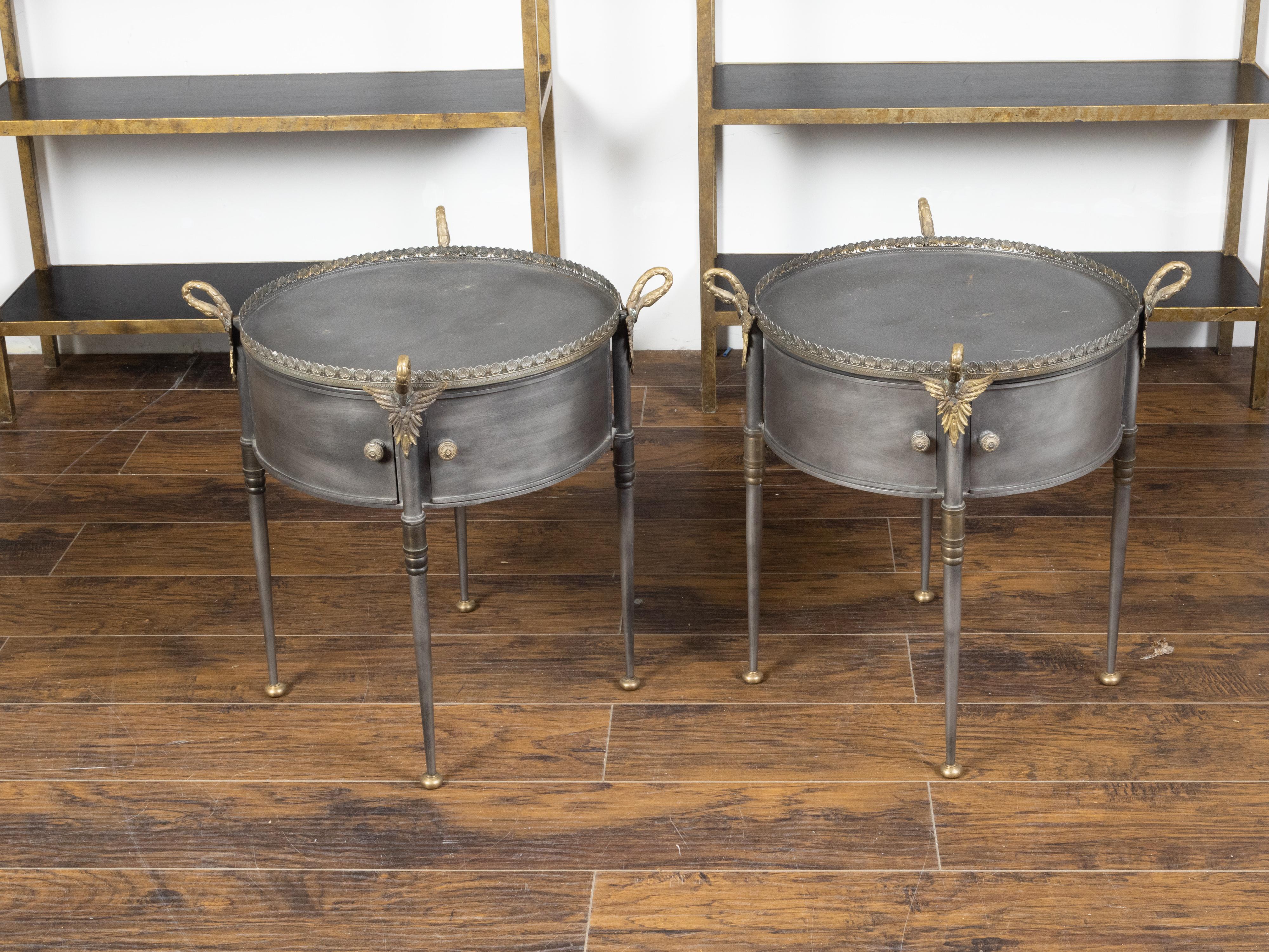 A pair of Trouvailles metal and brass round top side tables from the mid-20th century with two doors, swan necks and wing motifs. Created by the American company Trouvailles, each of this pair of metal side tables features an exquisite circular top,