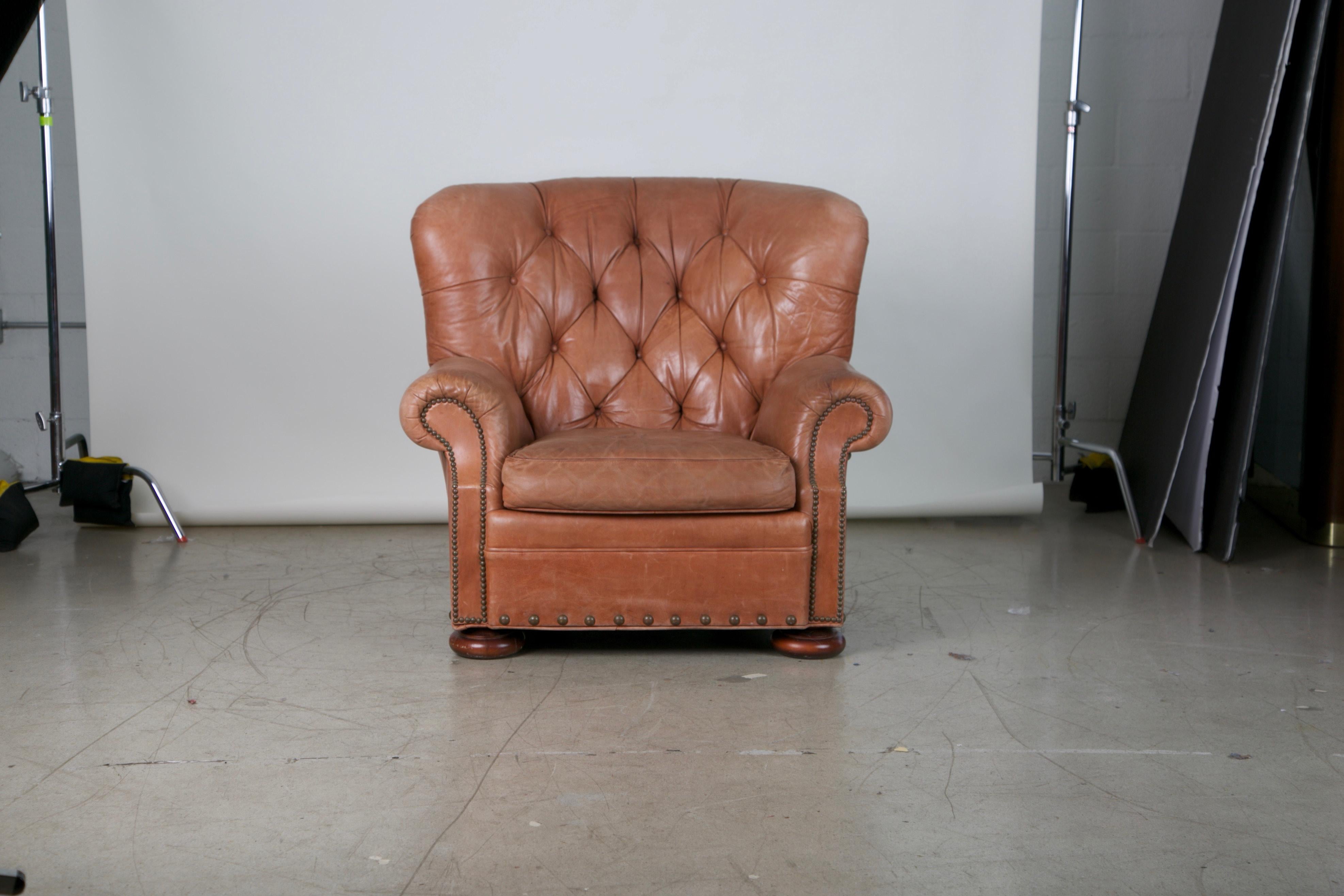 Handsome pair of lounge chairs in the style of Ralph Lauren. This set would inject some traditional elegance to any interior, featuring rolled arms, tufting, and sizable bronze nailheads. The duo exhibits the original tan leather upholstery that has
