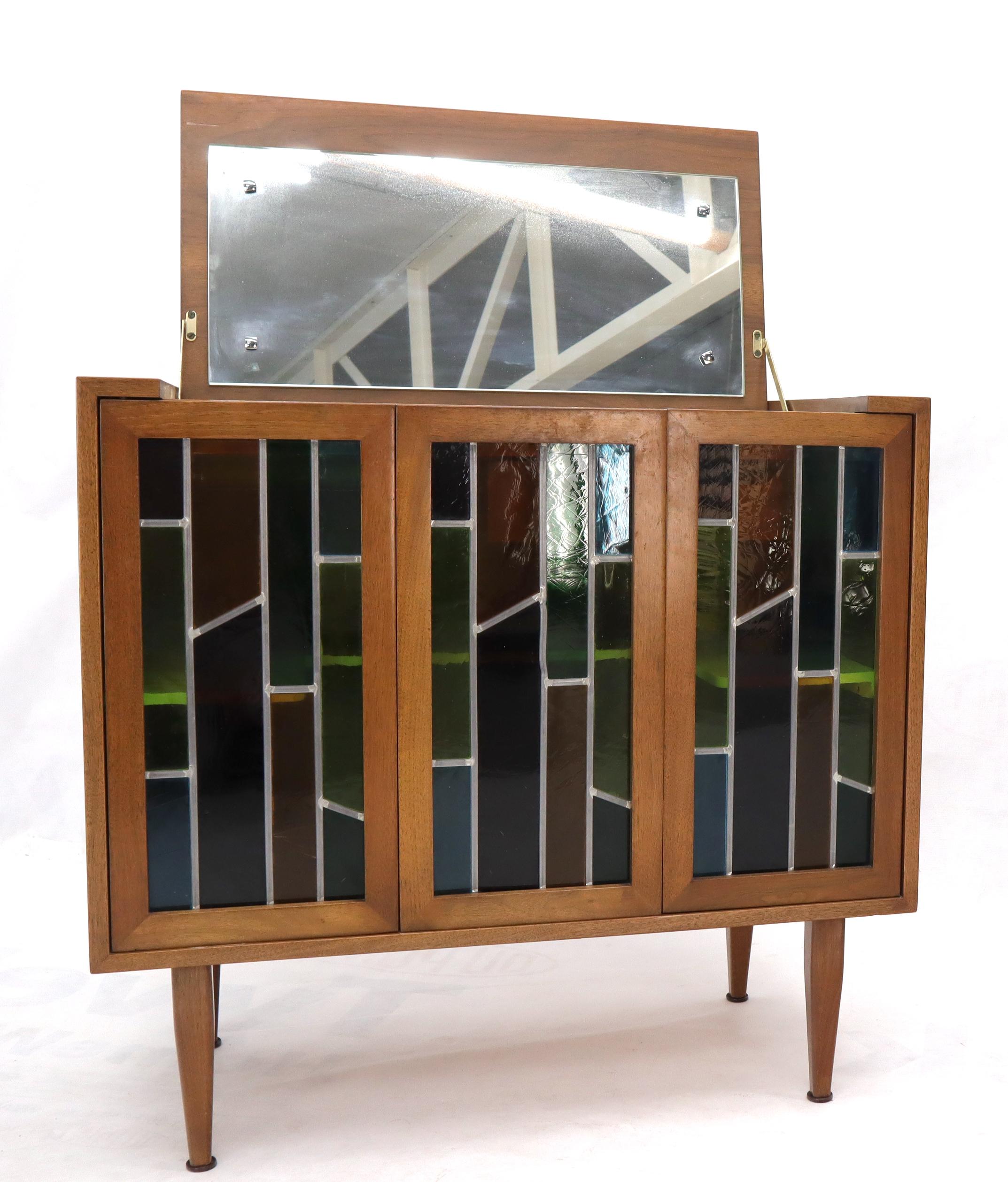 Pair of vintage Mid-Century Modern good quality walnut cabinets. The folding stained glass cabinet features lift mirrored top, brass hinges, internal lights and functions as liquor cabinet or bar. The second cabinet features folding doors and deep