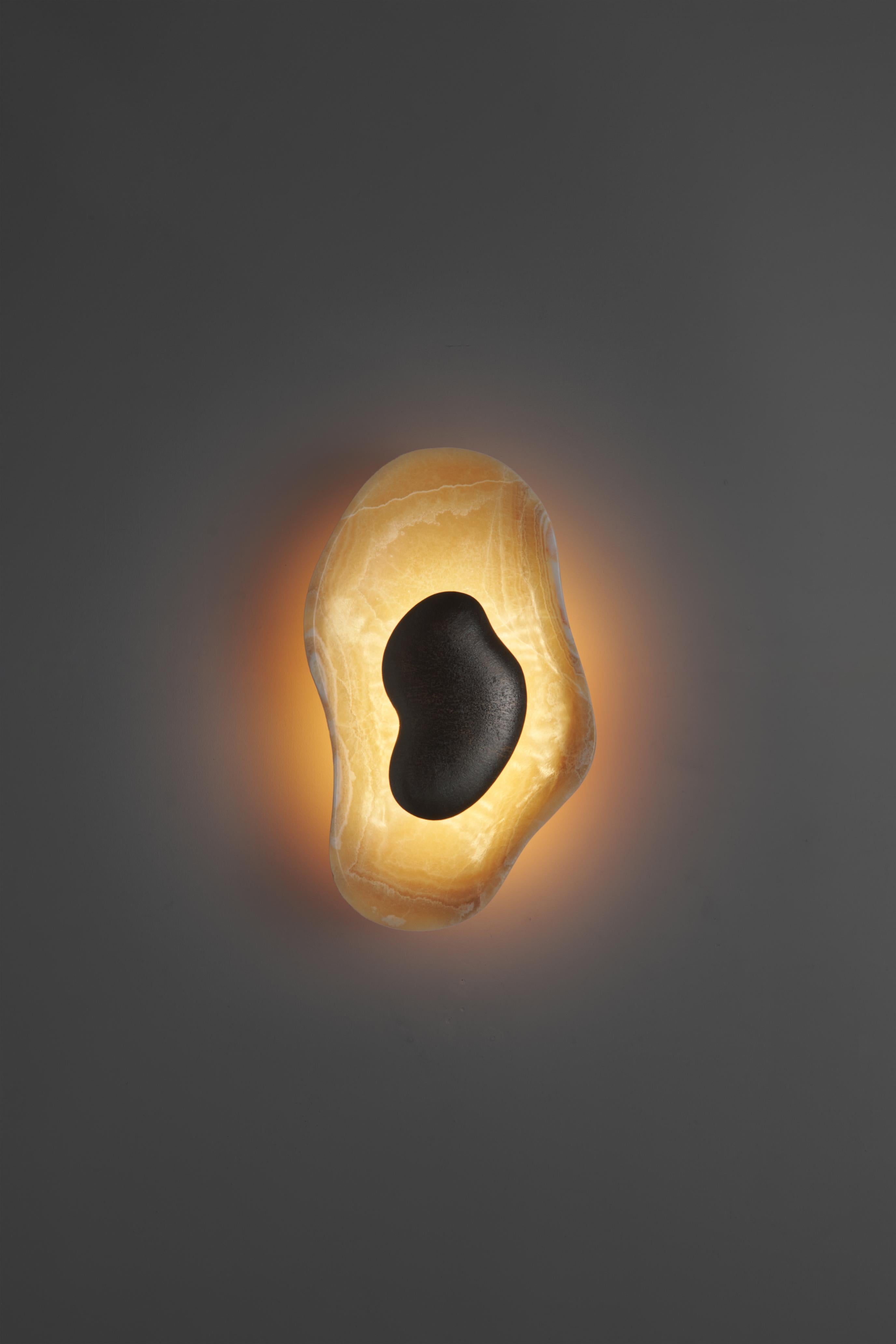 Two mirror-image organic forms, handcrafted from rare mustard-yellow onyx, with unique and unrepeatable veining. At the center, a hammered bronze element with a fluid, sinuous shape appears to float on the stone, diffusing light and enhancing its