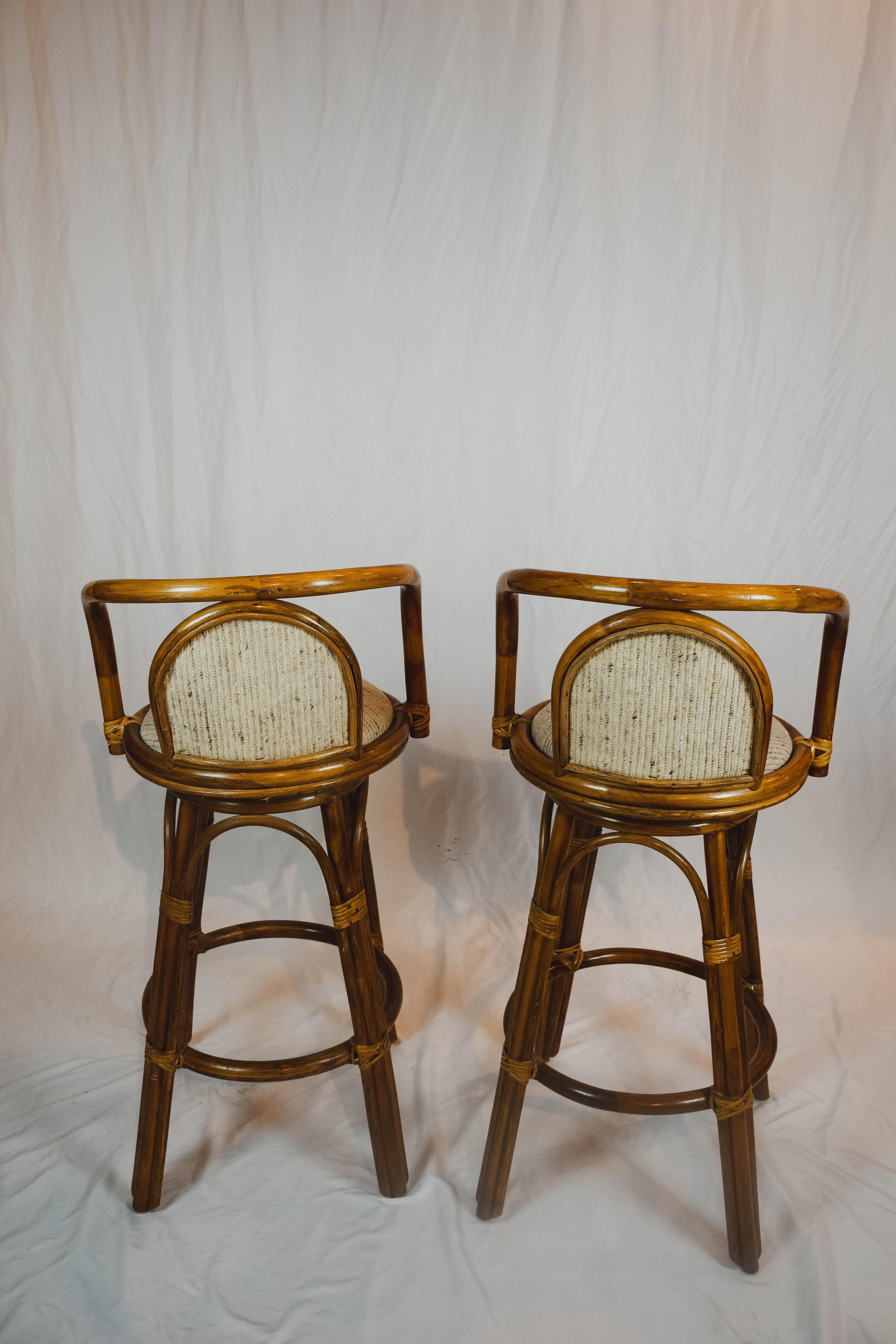 Pair of Tiki upholstered bamboo and rattan midcentury bar stools. These bentwood stools are bar height, with swivel seats and foot rests. They are upholstered in a neutral cream/beige fabric.