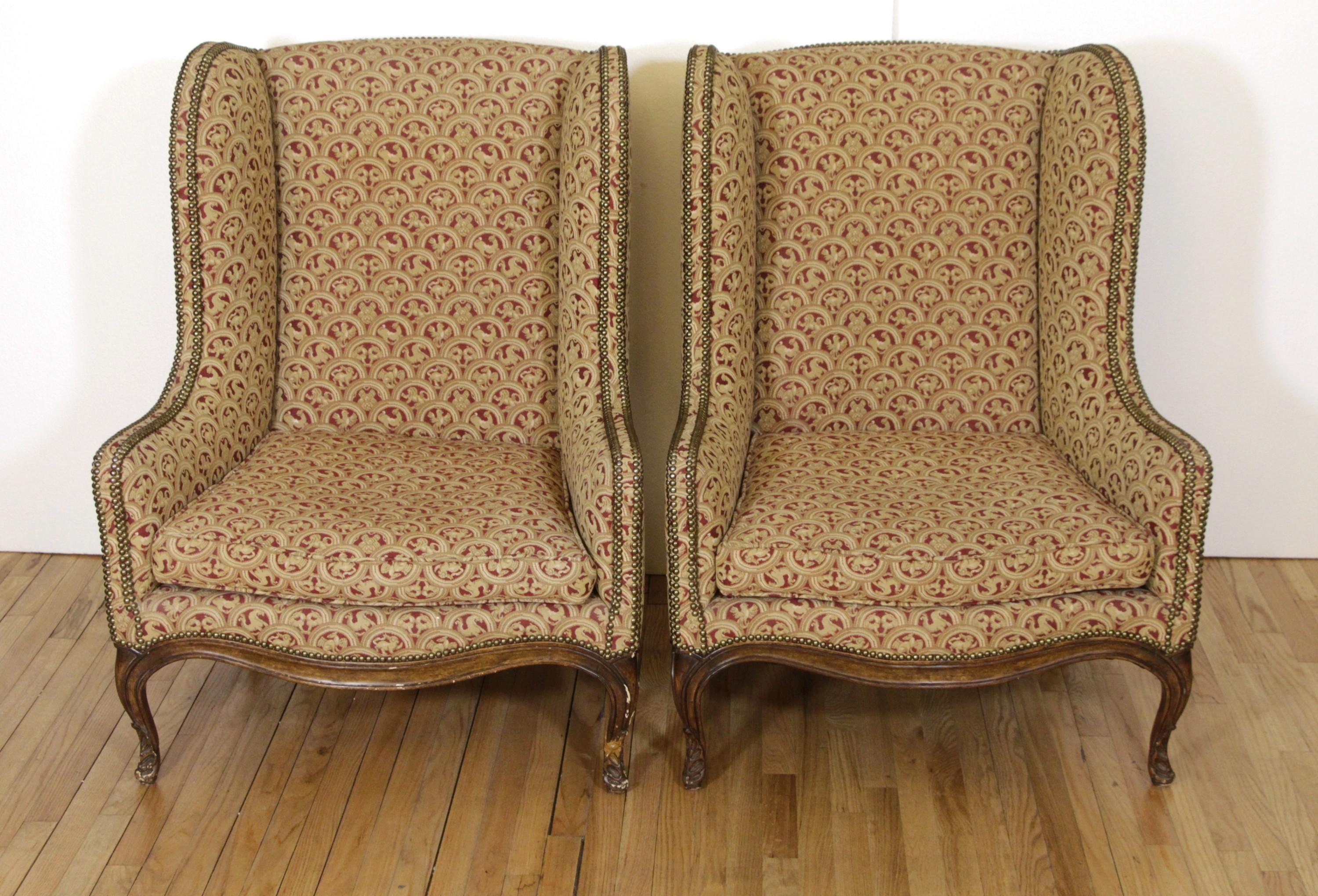 A pair of 20th century winged-back and upholstered armchairs with a finely carved wood base. The upholstery consists of semi-circle red and beige figural details. There are brass studs along the arms and sides. This can be seen at our 333 West 52nd