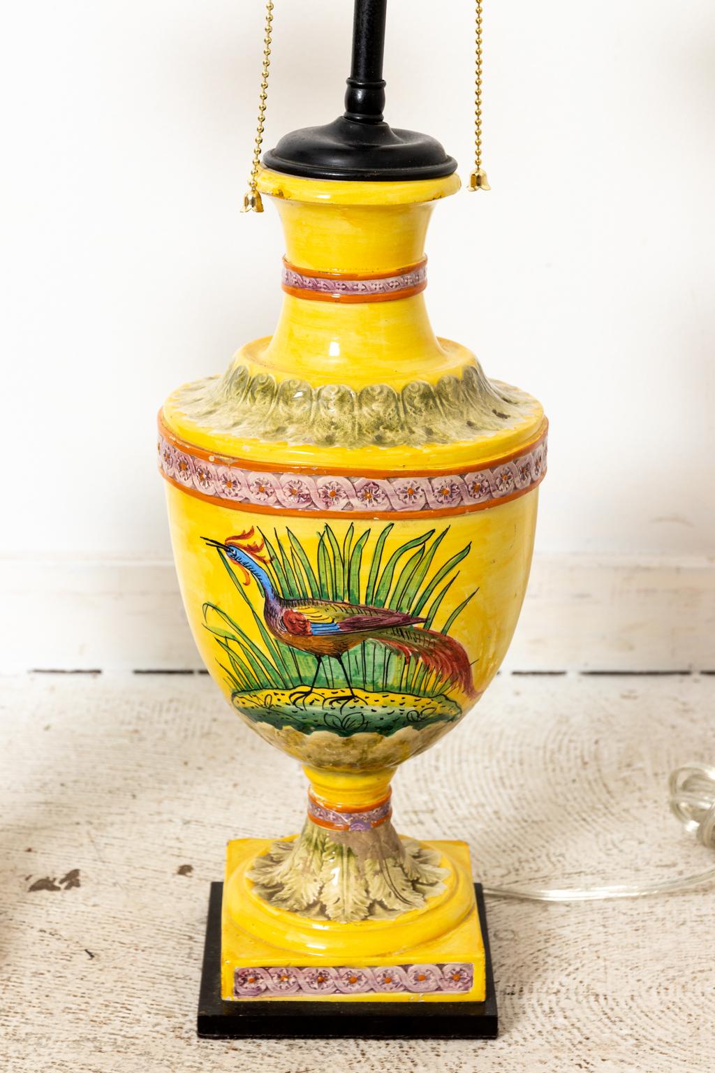 Circa 1910-1915 pair of urn form yellow glazed Majolica lamps with painted foliage and guilloche trim. The lamps are further illustrated by a large central panel of a multi-colored bird. Please note of wear consistent with age including repairs to