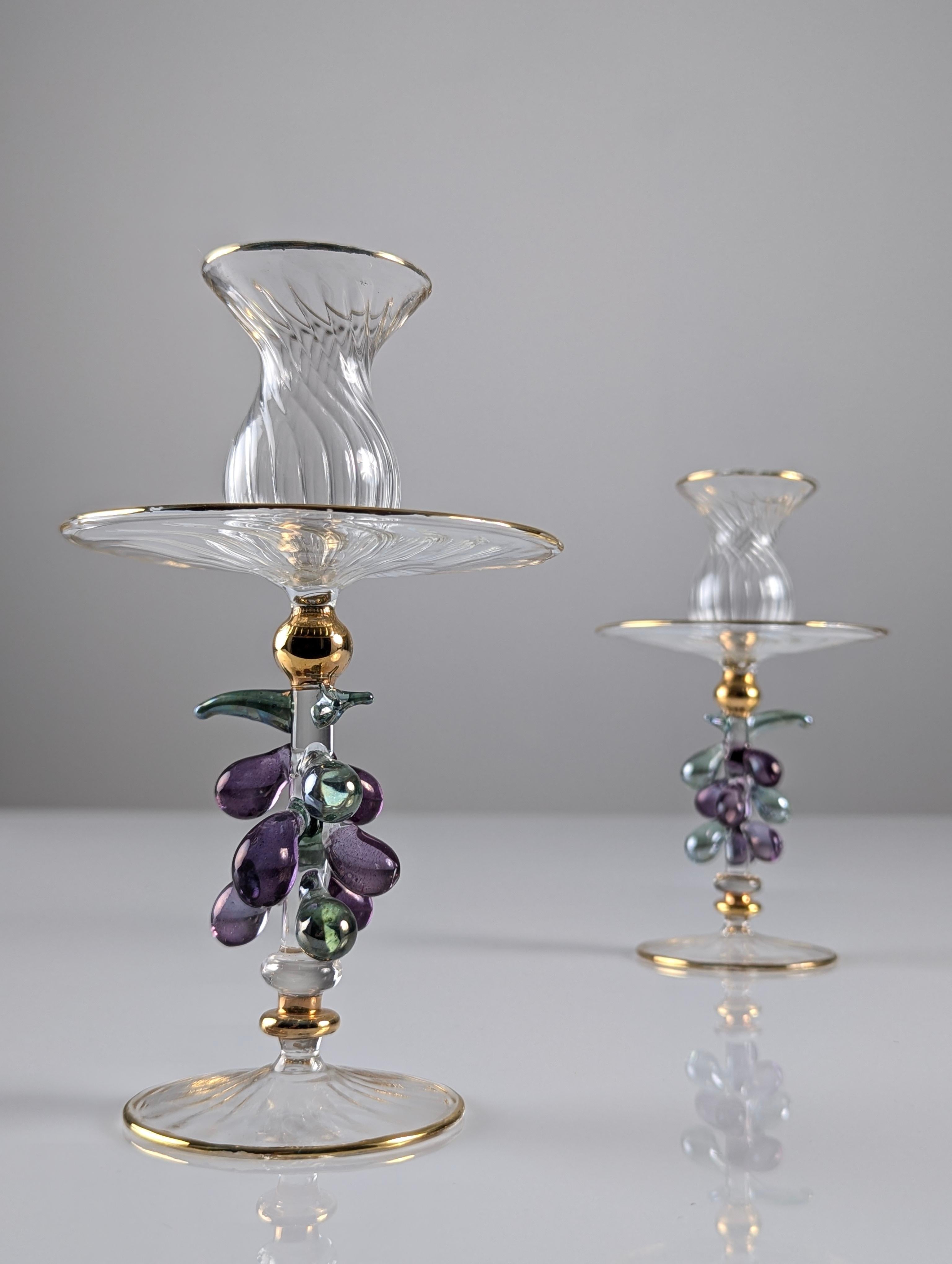 Questa elegante coppia di candelieri è realizzata in vetro veneziano con una squisita lavorazione a mano. I candelieri presentano delicate decorazioni sui bordi e sulla base superiore, evidenziate da una raffinata placcatura in oro, che accentua la