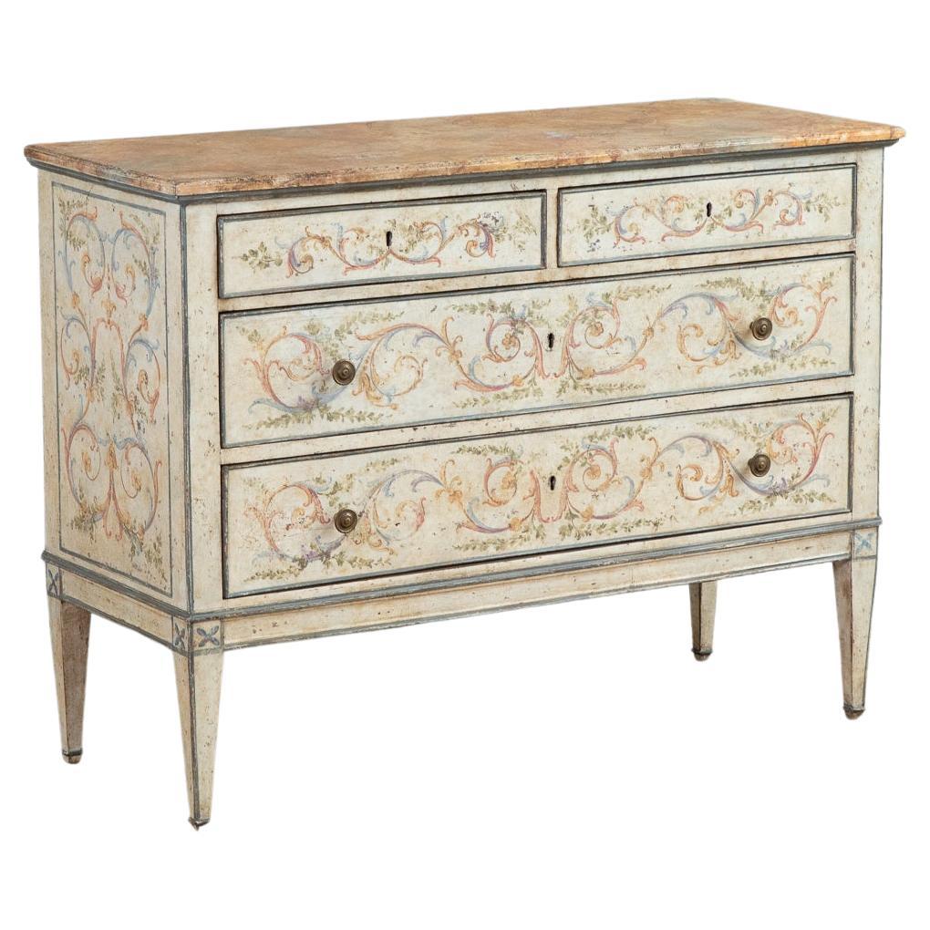 A pair of Venetian commodes, hand painted in the Neoclassical style featuring fine scroll work in hues of brick pinks, yellow ochres, olive greens and blues on an old white naturally aged, craquelure background. The overall warm tones of the