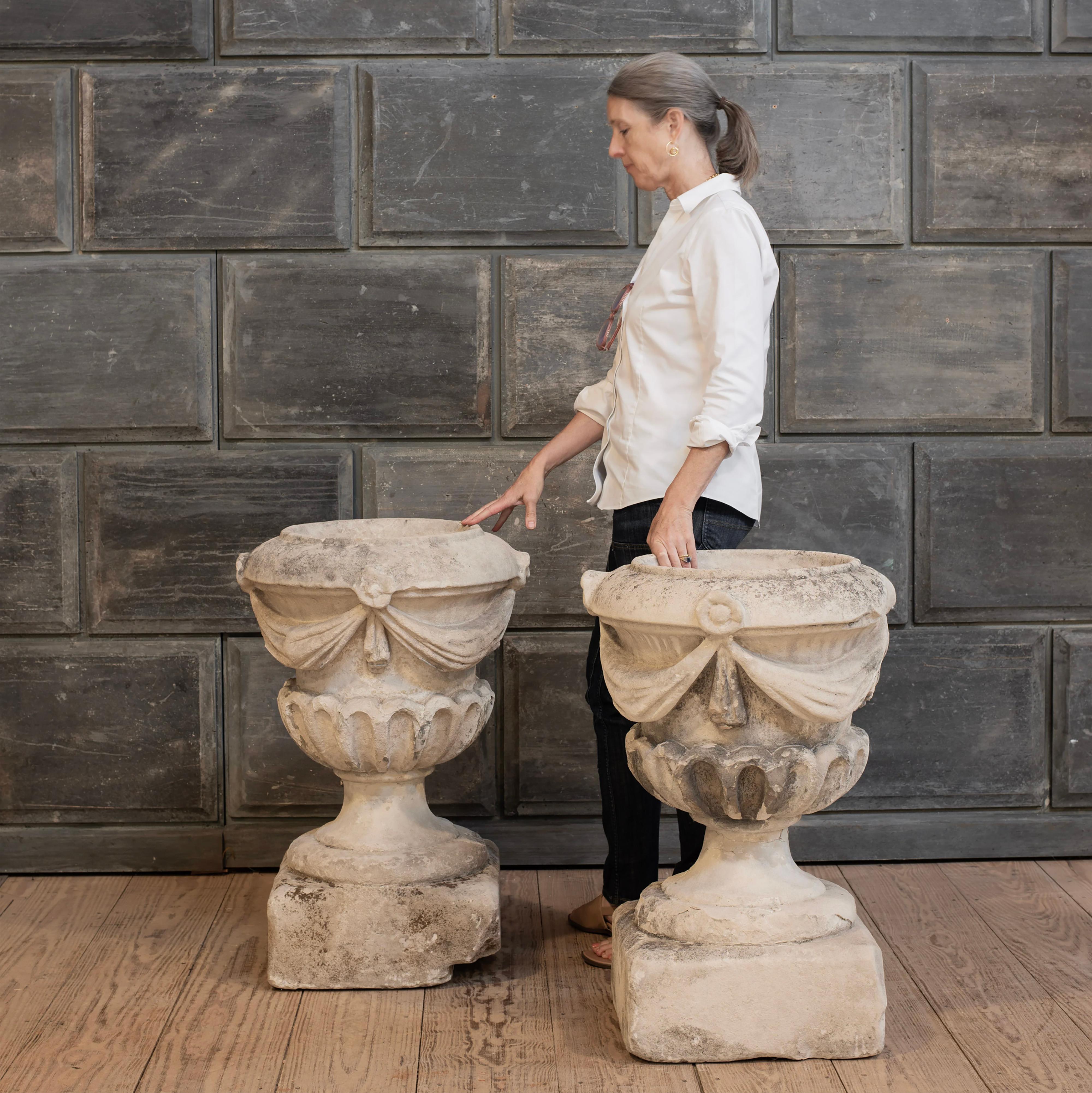 A large scale pair of very good 18th Century carved limestone garden urns featured stepped bases, draped swags, and flouted bowls. In great antique, true-to-age condition with some surface and material loss throughout. Structurally stable. One base