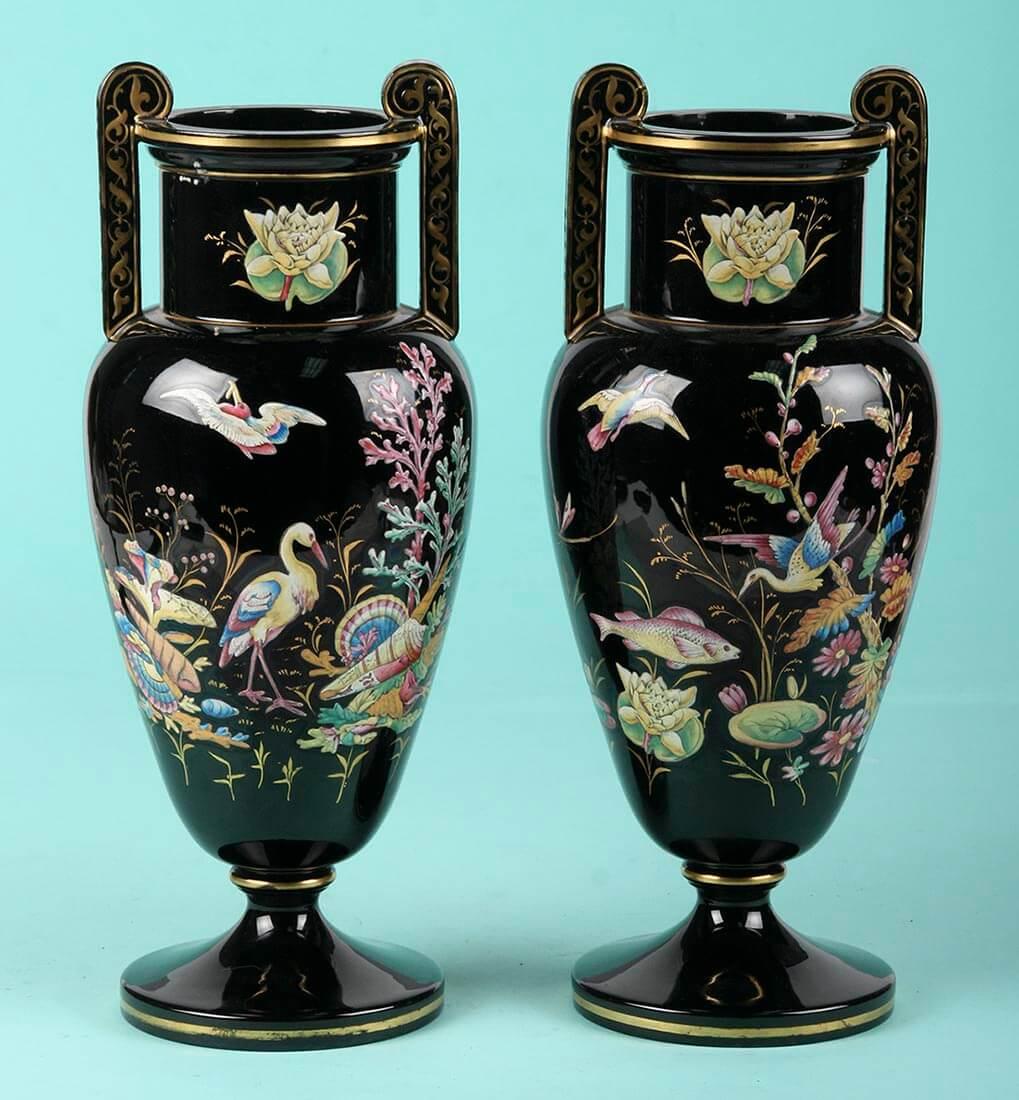 A pair of tall decorative Victorian vases, painted with enamel glass-paint.
The vases are made of porcelain or stoneware.
The black surface with colorful design is typical for the British Victorian style.
The gold decorations are painted by