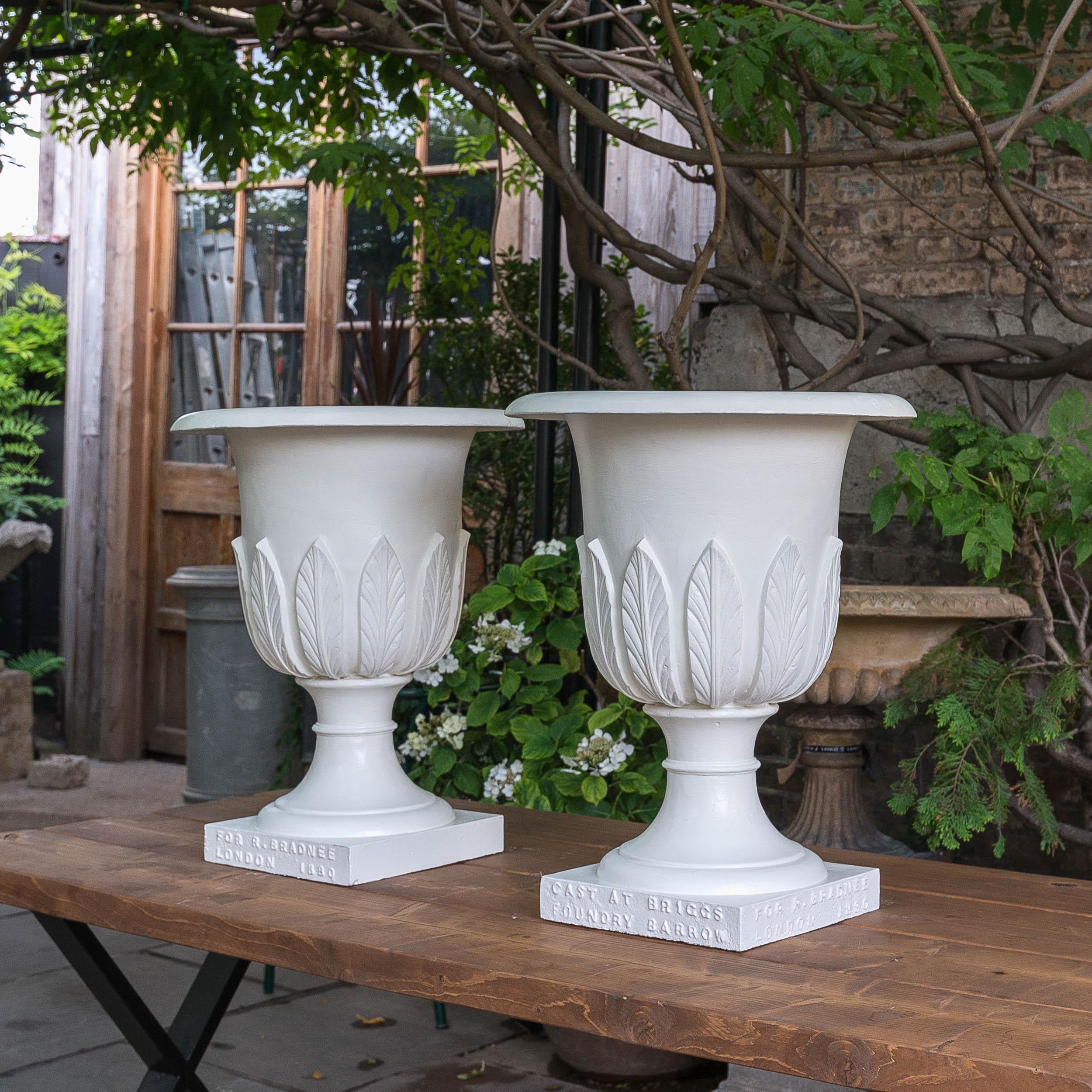 A pair of Victorian cast iron Campana urns, dated 1880, the body with stiff-leaf decoration, the plinth bases with embossed text, 'CAST AT BRIGGS FOUNDRY BARROW', 'FOR R.BRADNEE LONDON 1880'. Repainted.