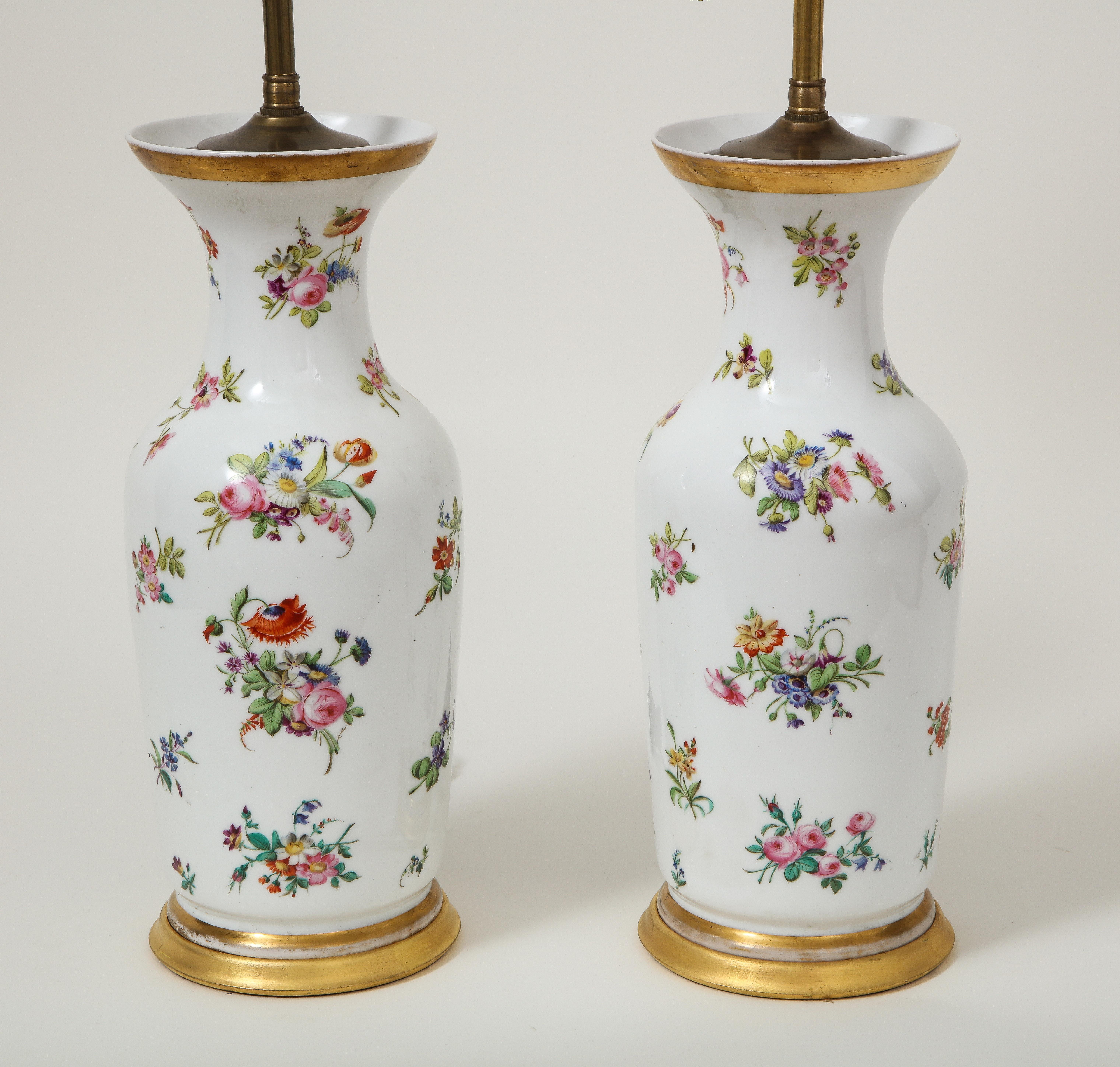 Each of baluster form decorated all-over with bunches of flowers, including auriculae, ranunculus, and forget-me-knots on the white porcelain ground; mounted on a turned giltwood base and fitted with adjustable brass rod and two bulb sockets.