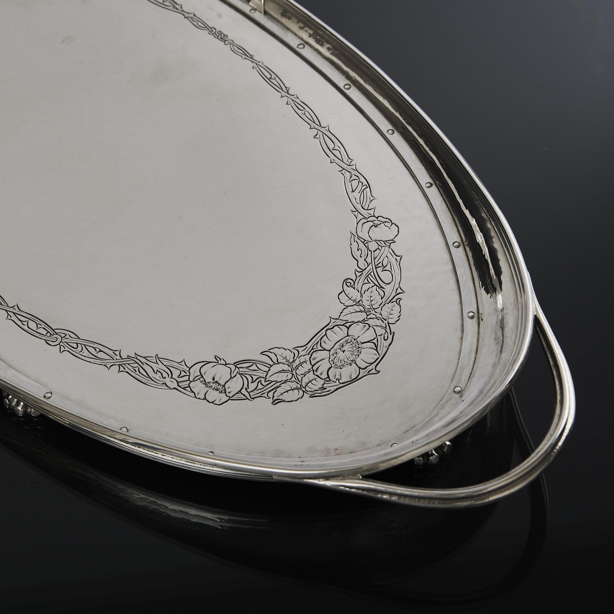 Made by Omar Ramsden, an Arts and Crafts two-handled navette-shaped silver tray, sitting on six paw feet and surrounded by a raised gallery. The body of the tray features spot-hand-hammered decoration and the centre is hand chased with a band of
