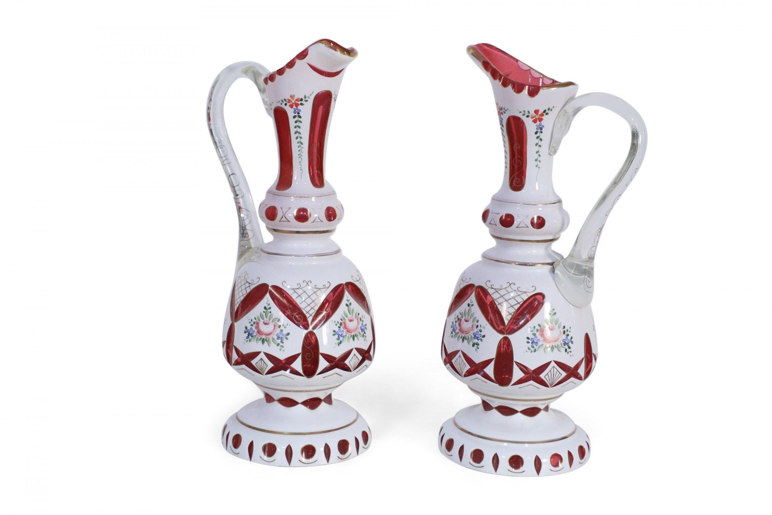 Pair of French Victorian white opaline overlay glass ewers / pitchers decorated with painted botanicals and gold motifs interspersed with transparent ruby shapes throughout and accented in exaggerated large glass handles (PRICED AS PAIR).
 