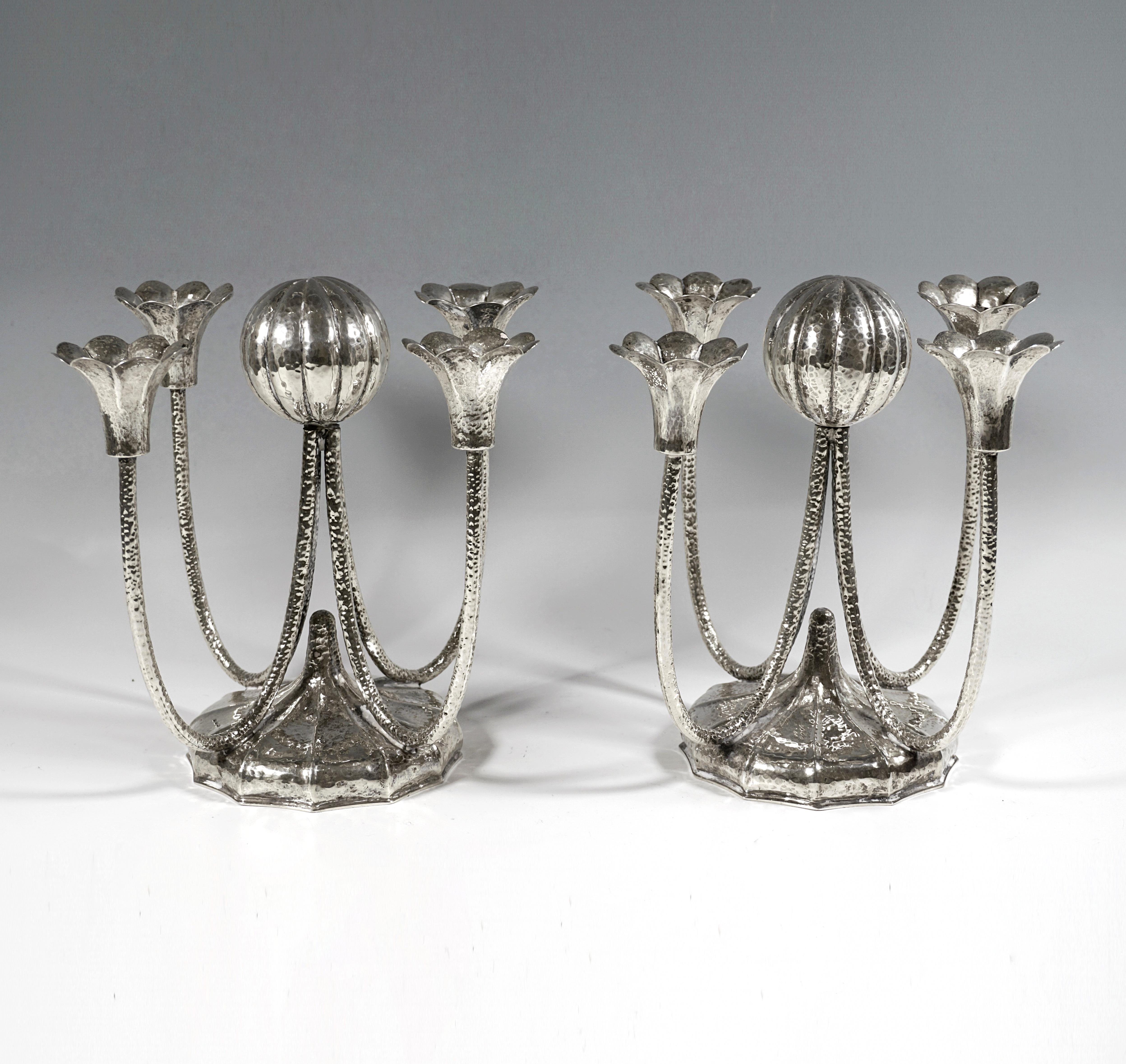 Two four-armed candleholders made of hammered and chased silver, base in a round basic shape, domed and ribbed and segmented in twelve rays, pointedly raised in the middle, with four slender U-shaped tubes on top, 
joined in the middle by a ball
