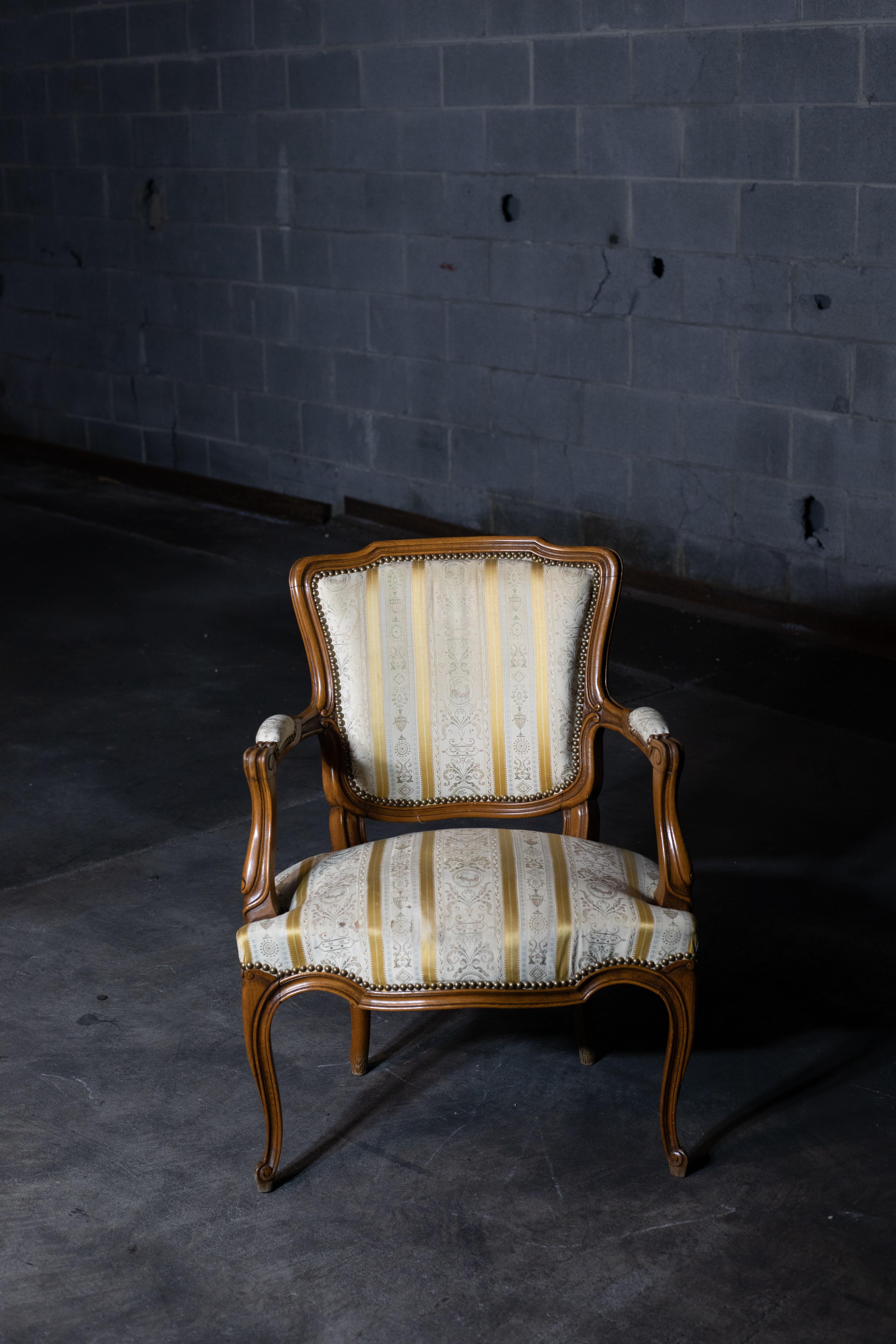 A pair of vintage armchairs in the style of Louis XV / Rococo. The chairs feature a carved wooden frame with cabriole legs and scrolling arms. Upholstered in striped cream and gold fabric with floral accents, finished with decorative nailhead trim.